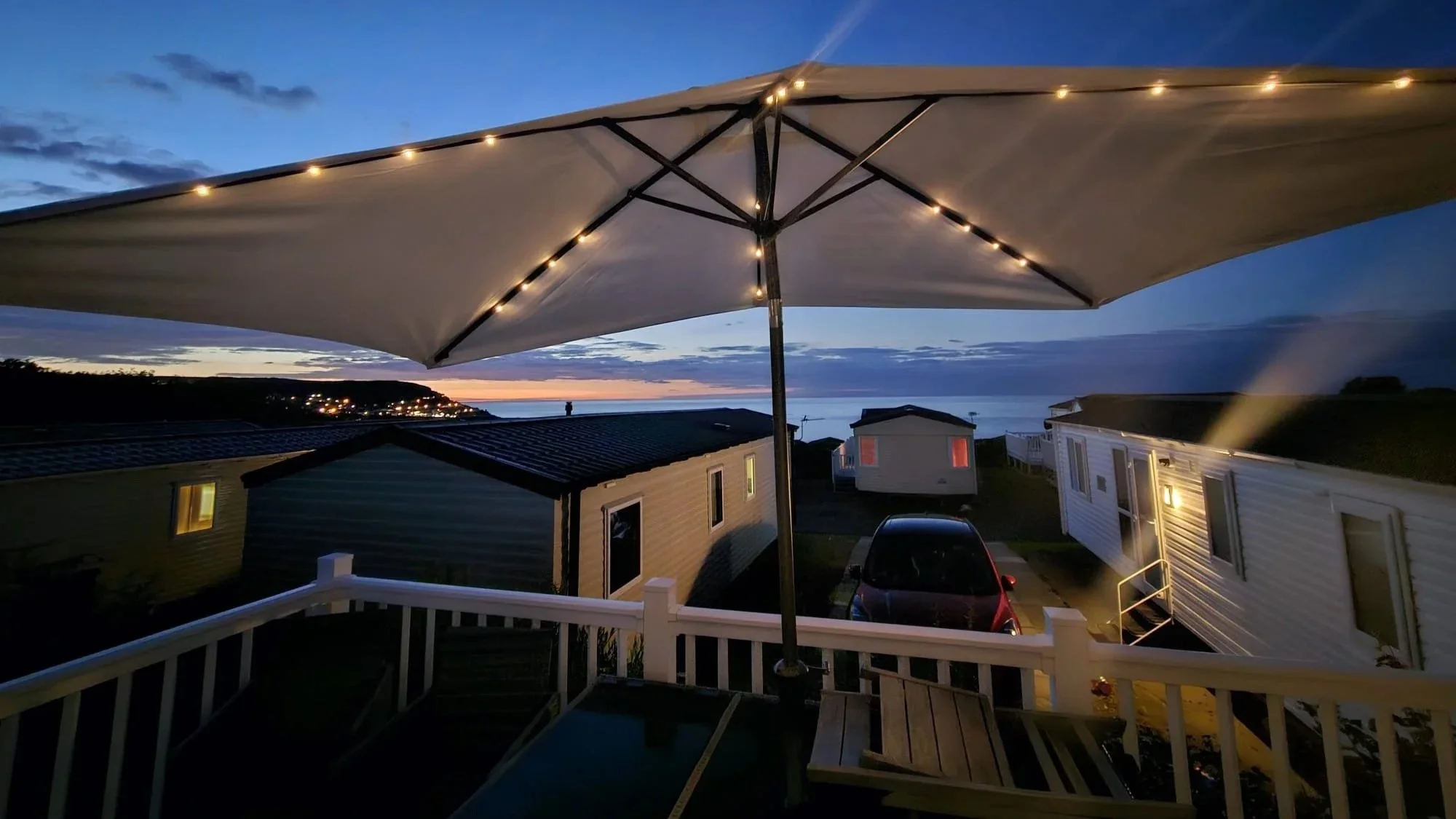 A view from a balcony showing a large umbrella with string lights, houses, a parked car, and a coastline at sunset or dusk.