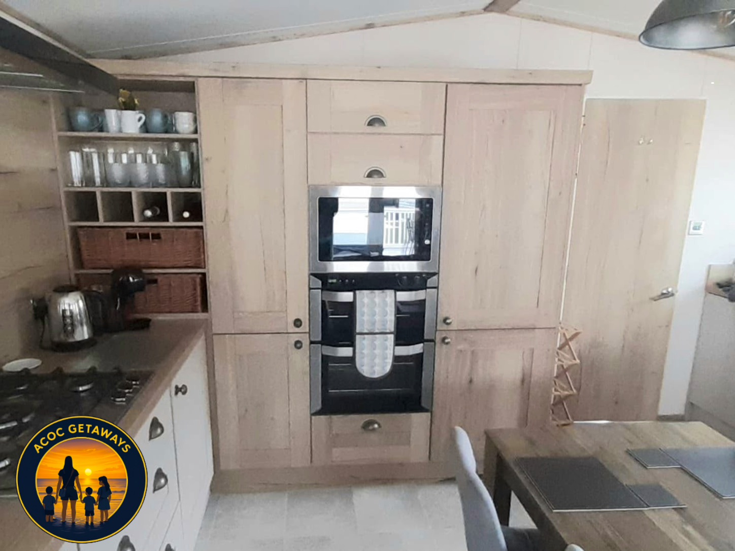 Kitchen with light wood cabinets, built-in oven, coffee maker, toaster, open shelving with cups and glasses, and a dining table with placemats.