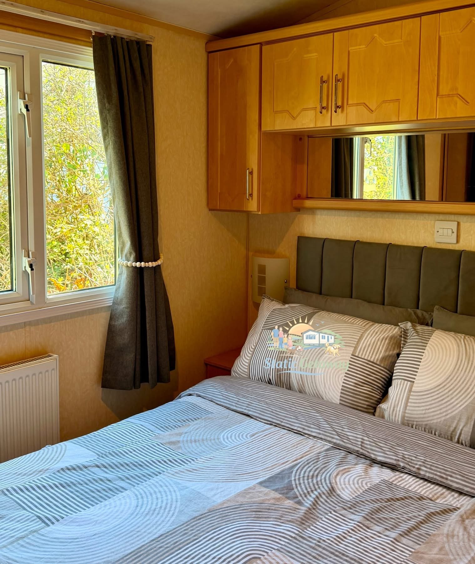 A cozy bedroom featuring a bed with striped beige and white bedding, a headboard with vertical gray panels, a window with dark curtains, wooden cabinetry above the bed, a small beige heater on the wall, and a view of trees outside the window.