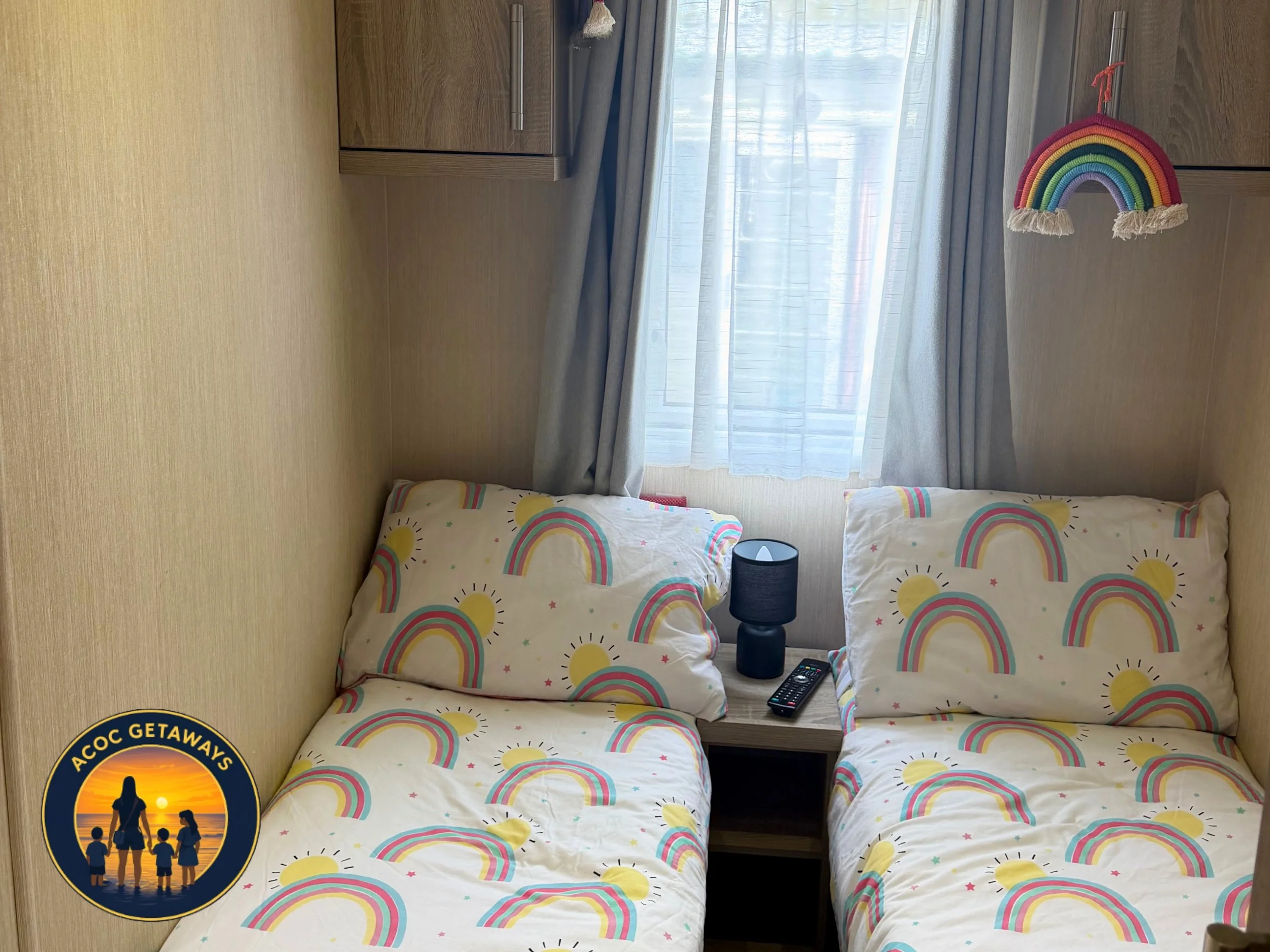A bedroom with beige walls, a window with light-colored curtains, and a bed with rainbow and sunshine patterned bedding. Two pillows with the same pattern are on the bed. There is a nightstand with a black lamp and a remote control. Two wooden cabine