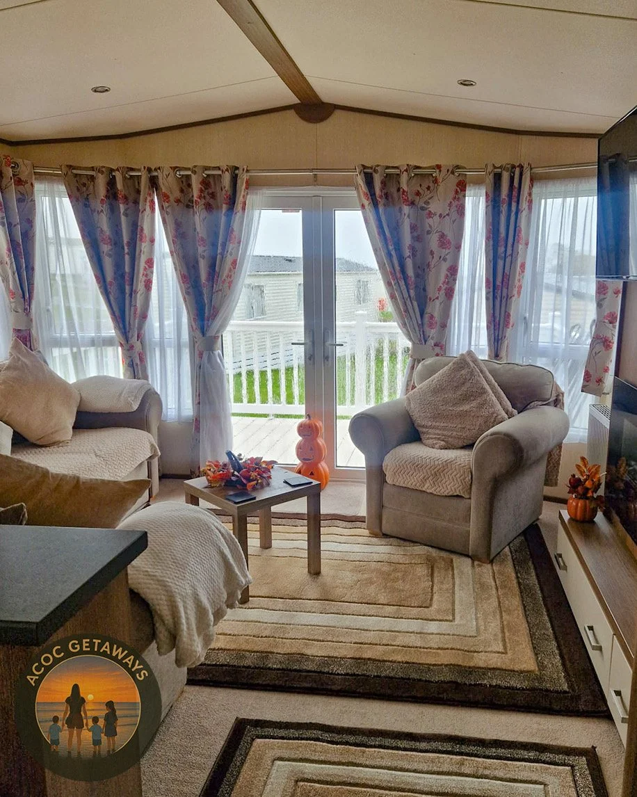 A cozy living room with large windows and floral curtains, featuring gray armchairs, a small wooden table, and a view of a porch with a green lawn outside.