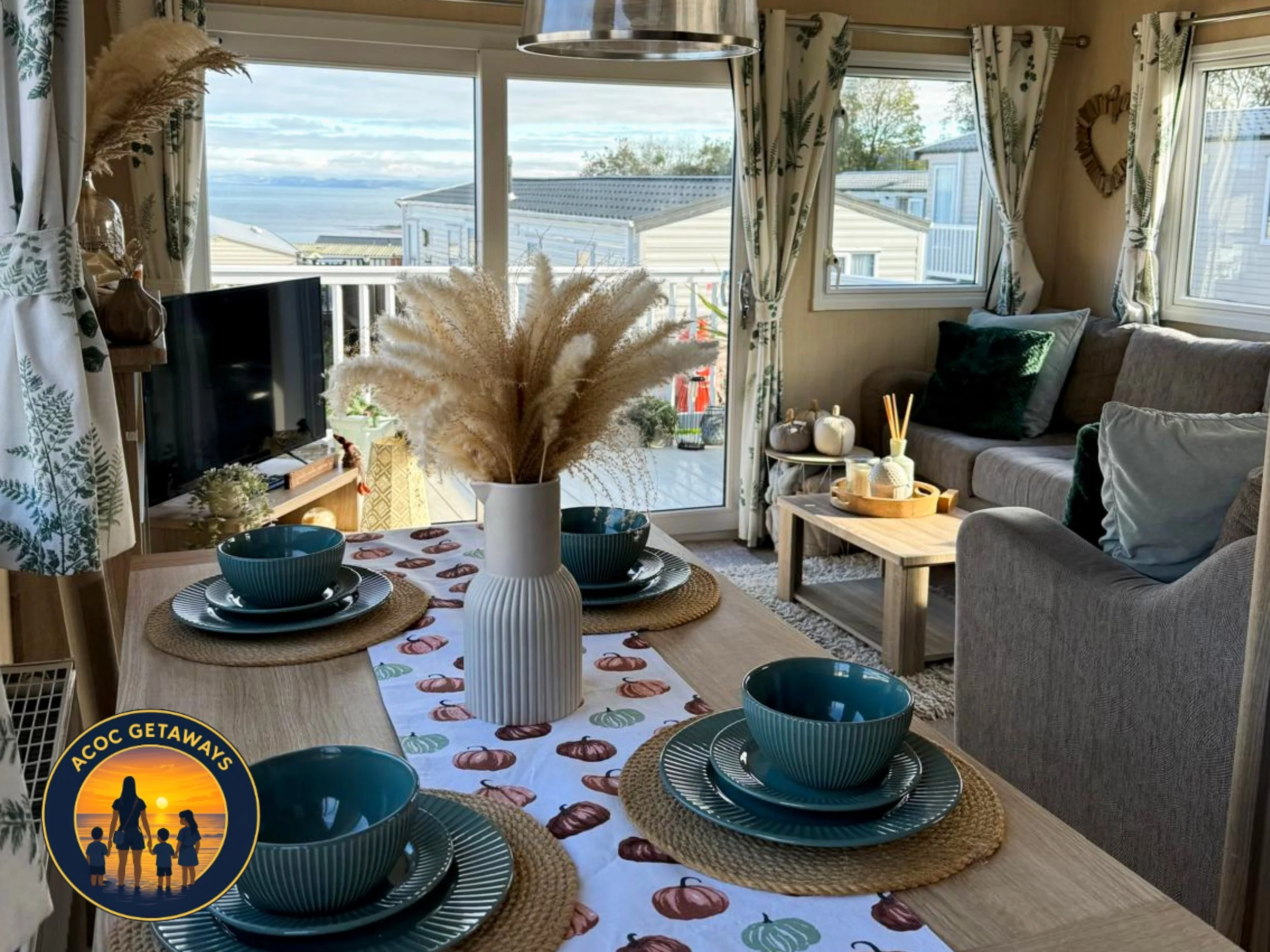 A cozy living room with large windows revealing a view of the ocean and sky, decorated with beige curtains patterned with green leaves. The room has a gray sofa with pillows, a wooden coffee table, and a dining table set with round placemats, a white