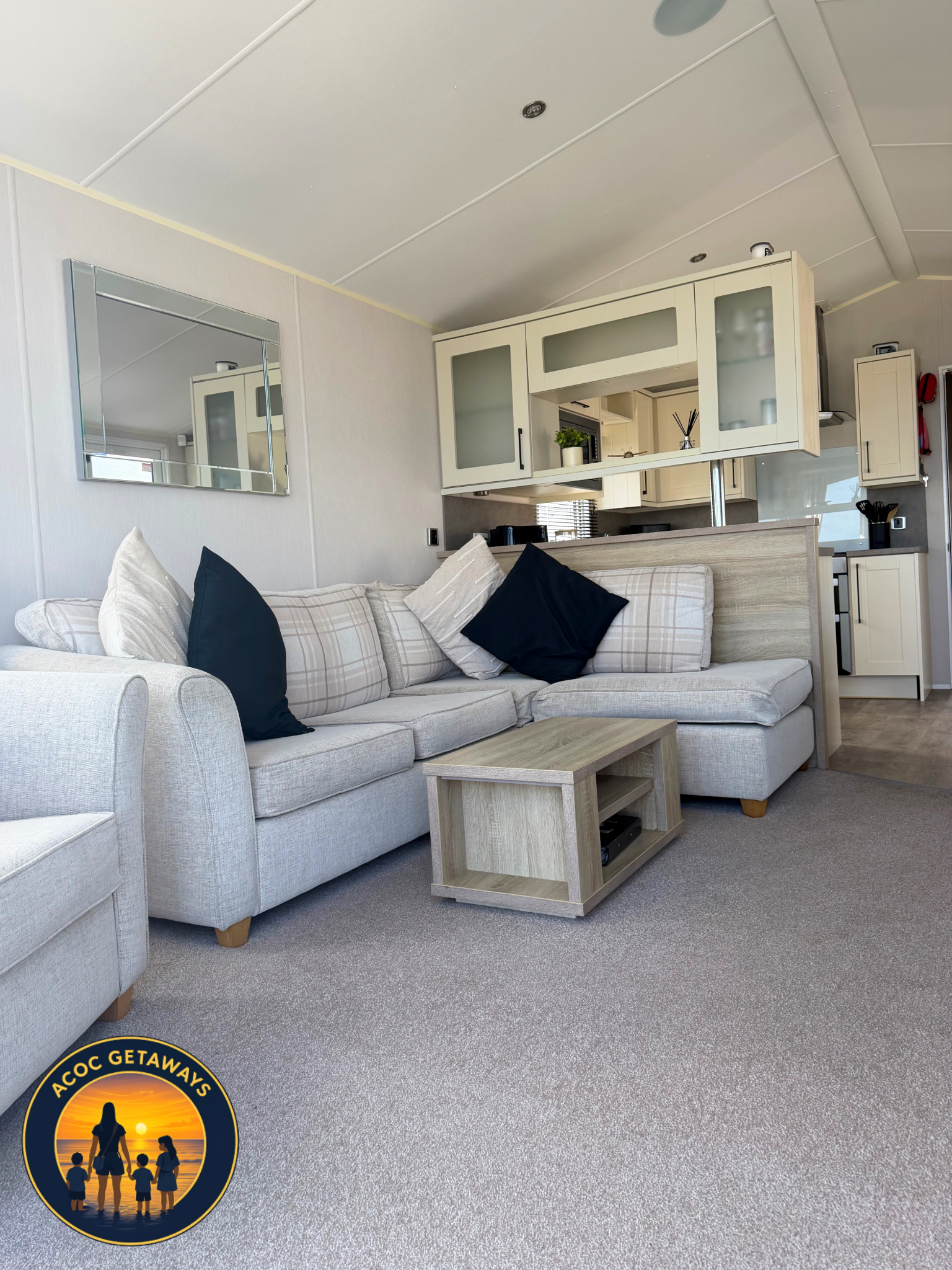 Living room with beige sofa, pillows, wooden coffee table, kitchen area in background with cabinets, plants, and decorative items, wall mirror, carpeted floor.