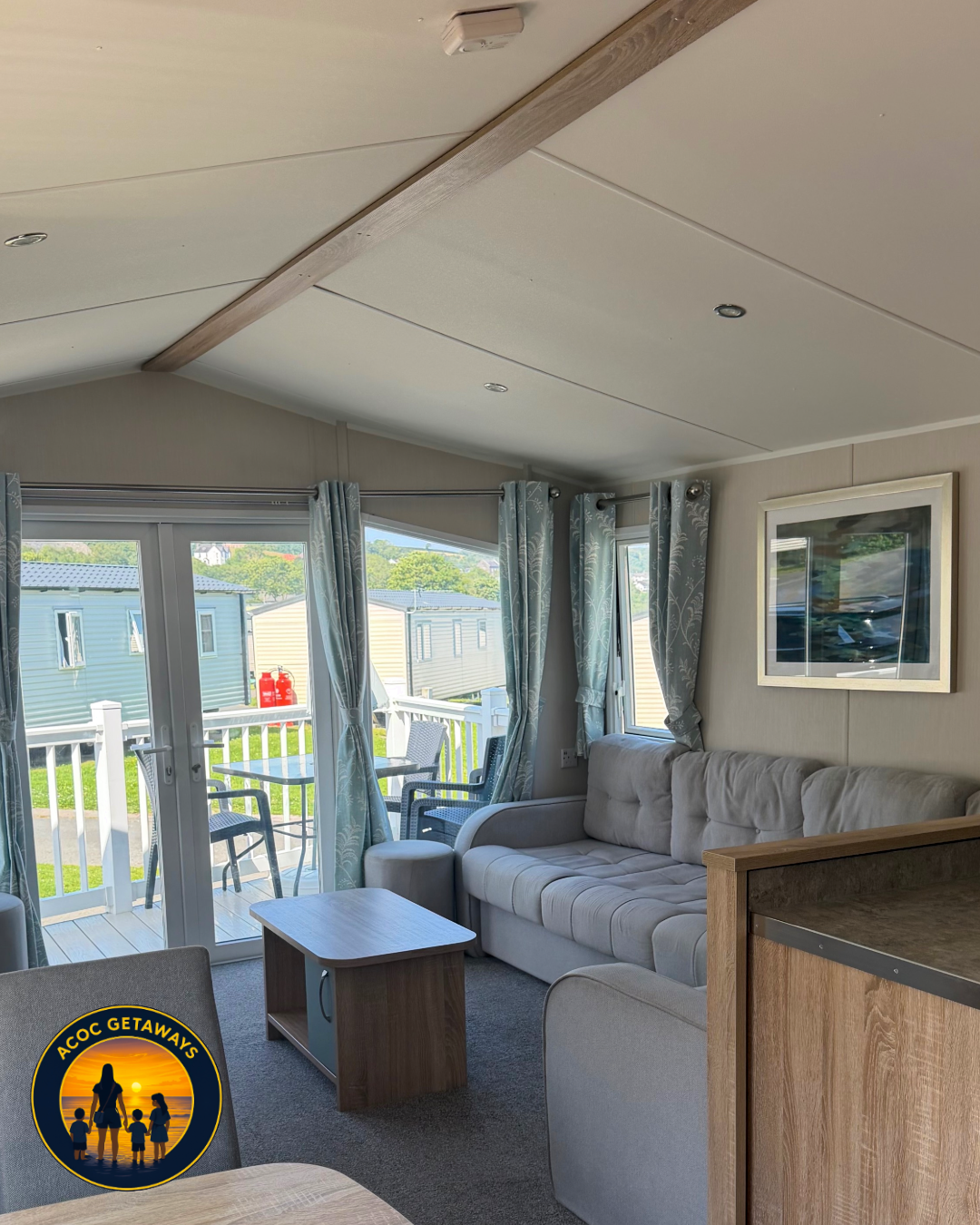 Living room with cream-colored sofa, coffee table, and sliding glass door leading to a balcony with outdoor furniture, surrounded by green trees and static mobile homes.