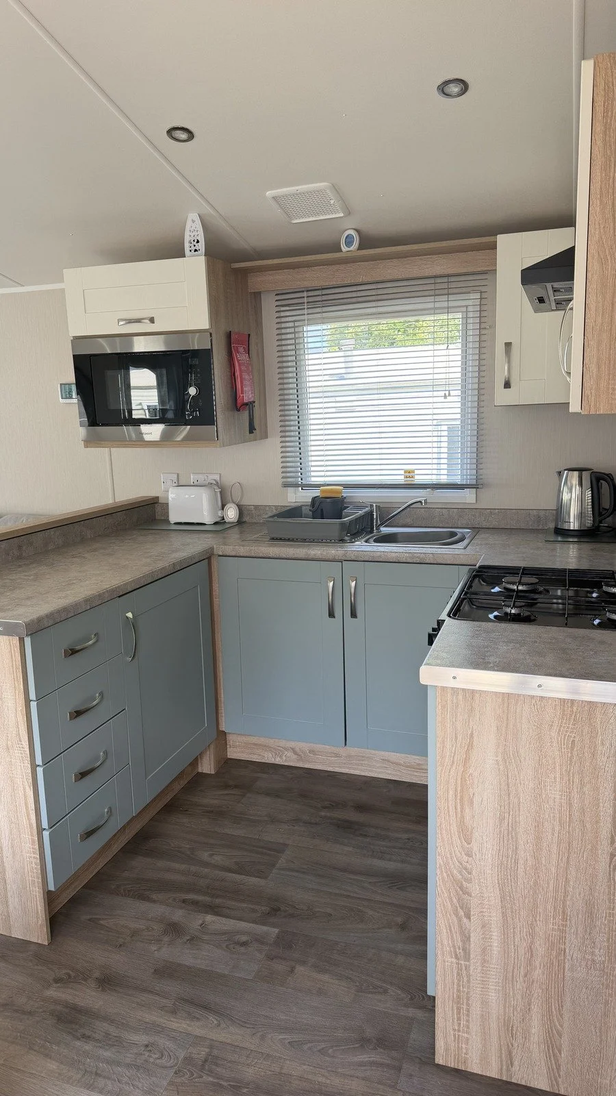 A compact kitchen with blue cabinets, a gray countertop, a window with blinds, a microwave oven, a toaster, a stove, a kettle, and a sink, with a wall and ceiling in neutral tones.