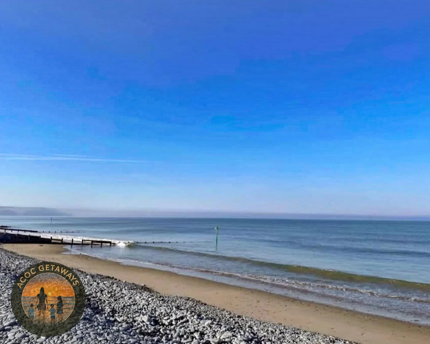 A peaceful beach with a pebble shoreline, calm ocean waters, a small pier extending into the water, and a clear blue sky. There is a logo in the bottom left corner that says "ACOC GETAWAYS" with an illustration of a woman and children.