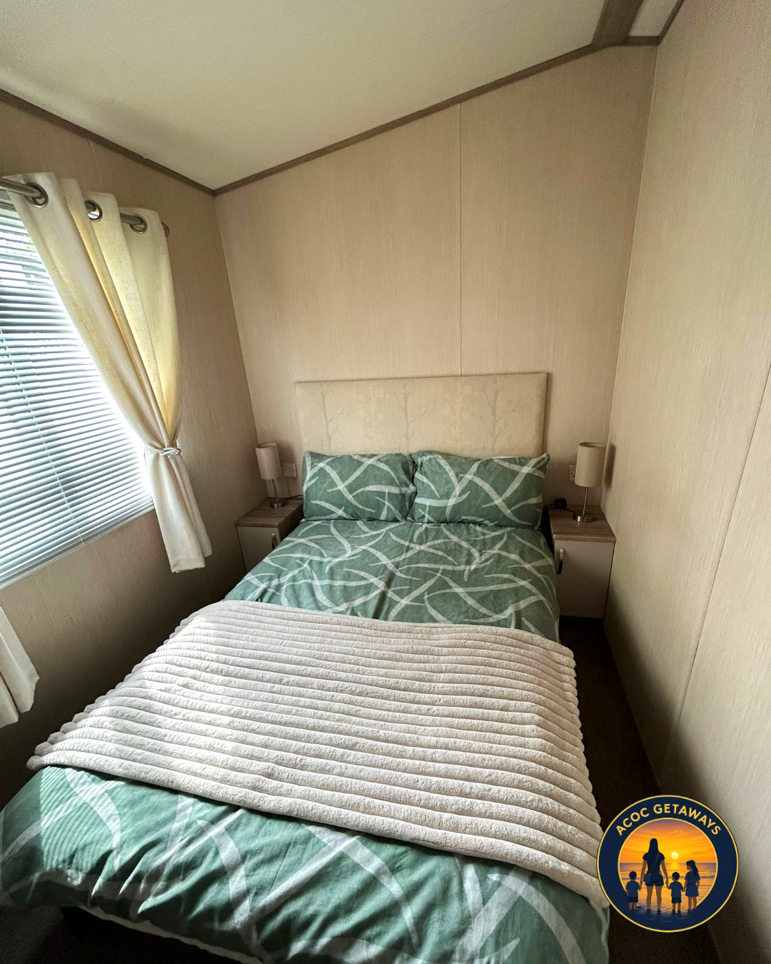 Small bedroom with a bed made up with green and white patterned bedding, beige headboard, two small nightstands with lamps, window with blinds and cream curtains, and dark flooring.