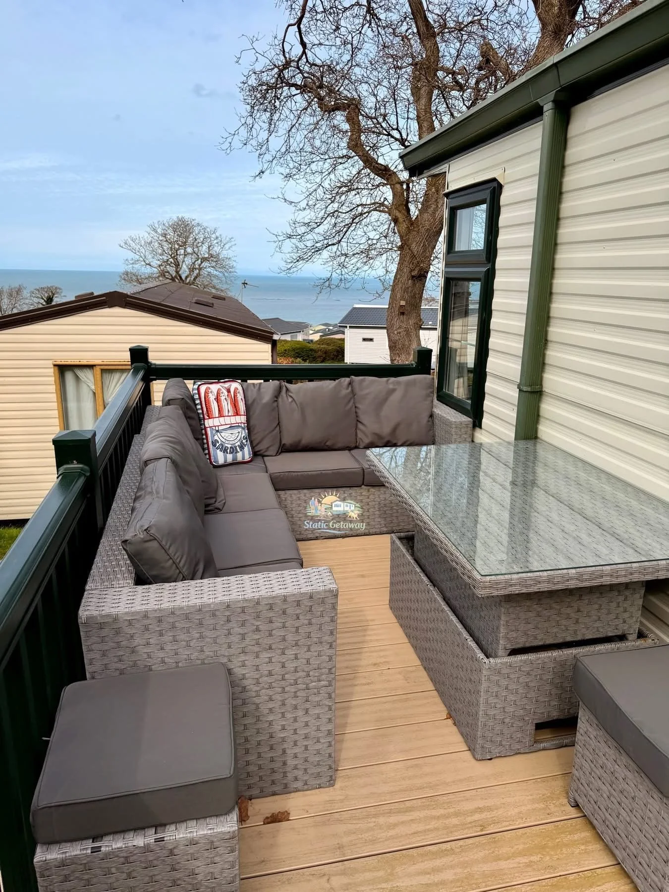 A balcony with a wicker sectional sofa, a glass-top table, and a small cushioned stool outside, overlooking houses, trees, and a body of water in the distance under a cloudy sky.