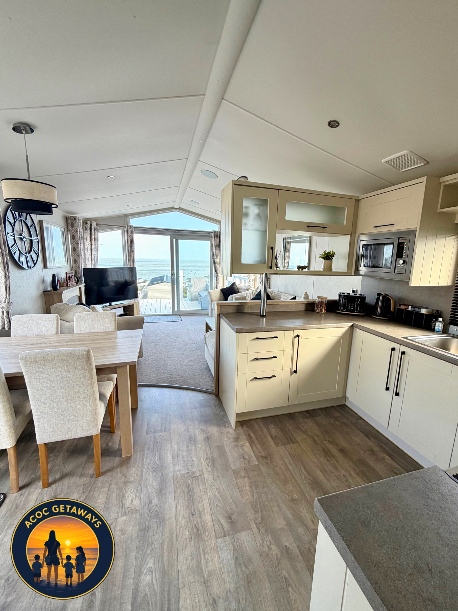 Interior of a modern living space with a kitchen to the right and a living room in the background. The living room features a TV, a coffee table, a sofa, and large windows with curtains showing an ocean view. The kitchen has cream cabinets, a microwa