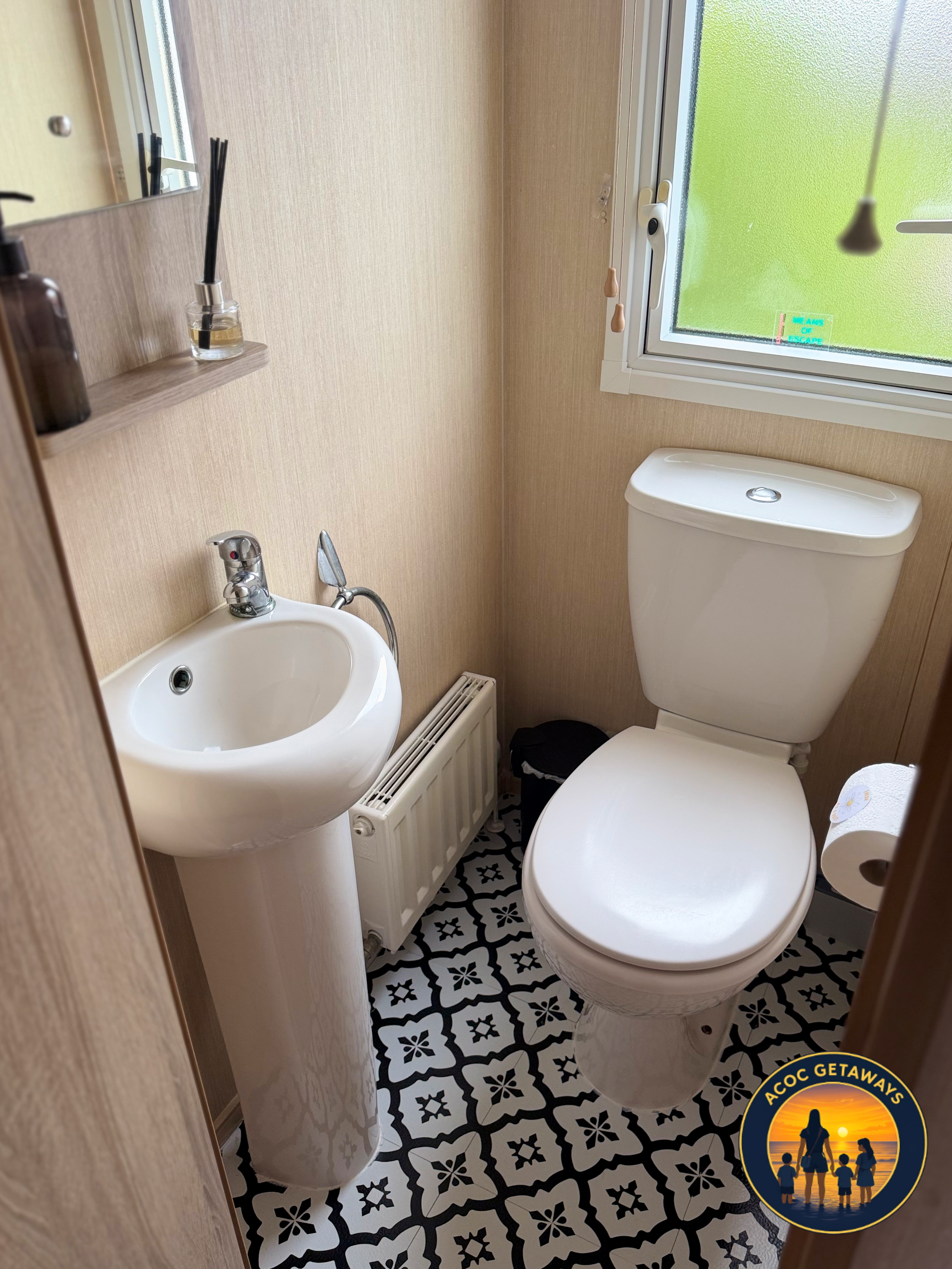 A small bathroom with a white toilet, a white sink, and patterned black and white floor tiles. There is a window with frosted glass, a small radiator below the sink, and a mirror above the sink. A small shelf holds a brown bottle and a diffuser. A ro