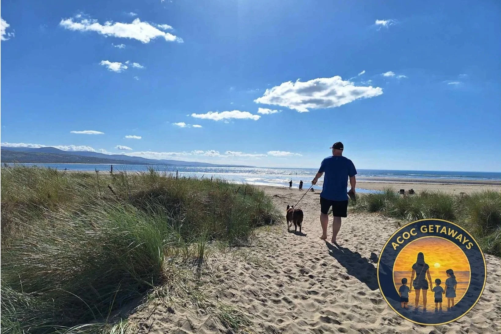 A man walking a dog on a sandy beach with grass dunes, ocean, and blue sky with clouds in the background. Several people are in the distance near the shoreline.