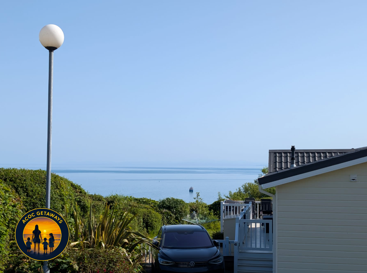 A coastal residential area with a view of the ocean, a boat, a house with a deck, a parked Volkswagen car, a street lamp, and lush greenery.