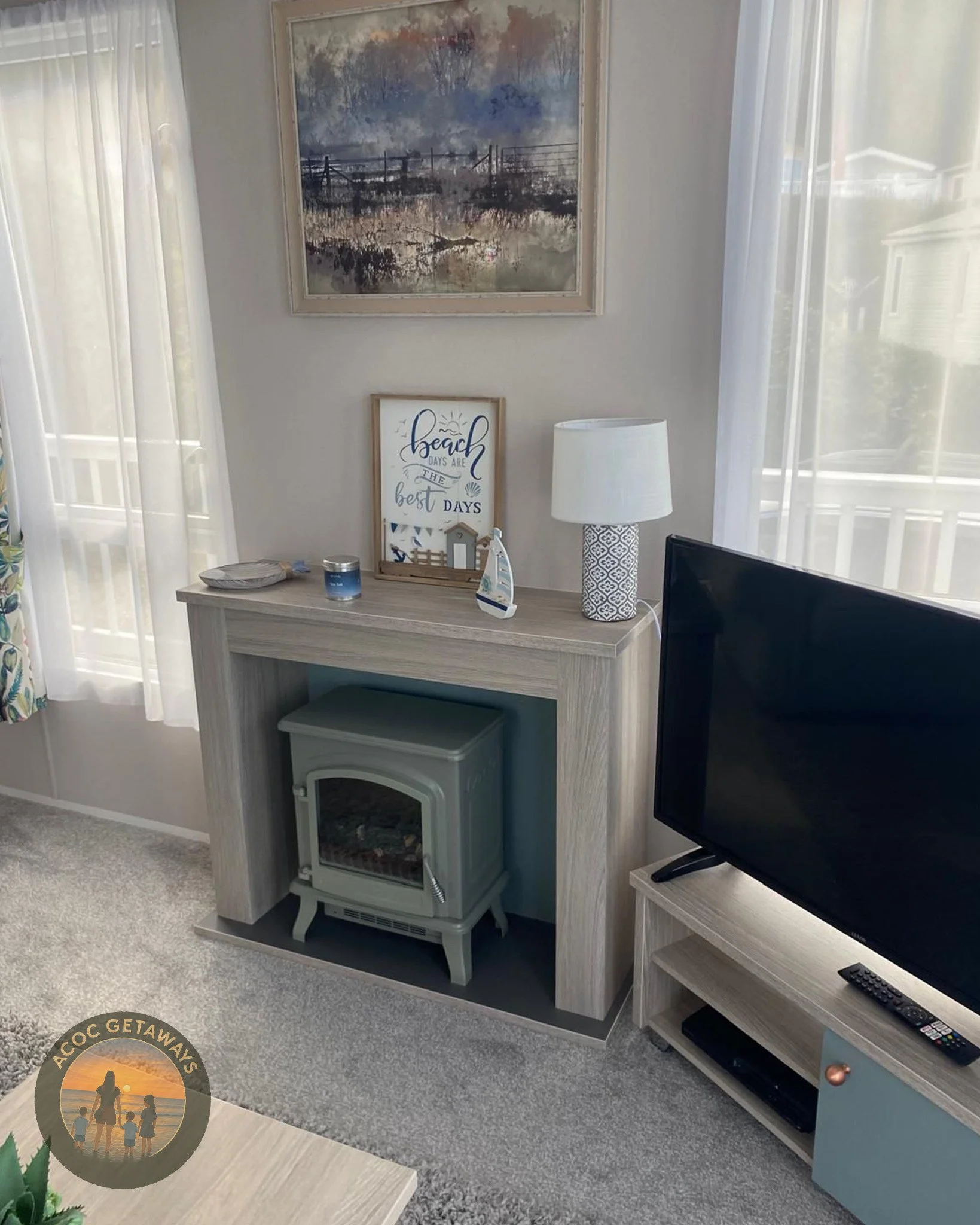 Living room corner with a flat-screen TV on a wooden stand, a light-colored fireplace heater underneath a mantle, a white table lamp, decorative items, and wall artwork. Light curtains on windows allow natural light.