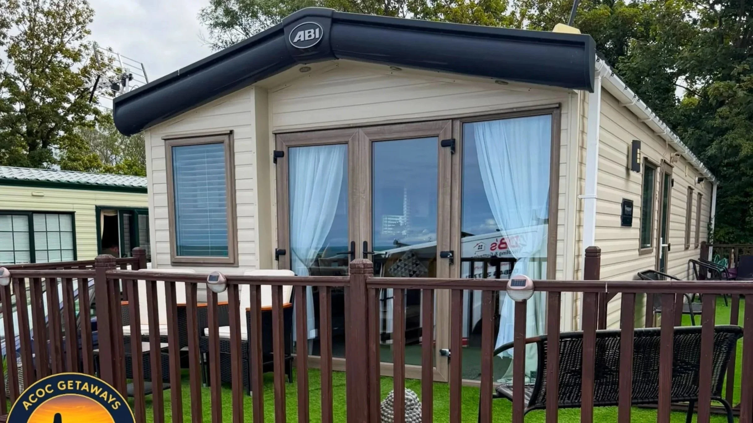 A small beige mobile home with brown trim, a large sliding glass door with curtains, and several windows. It has a black awning and is surrounded by a wooden deck and railing with outdoor furniture. There are trees and power lines in the background.