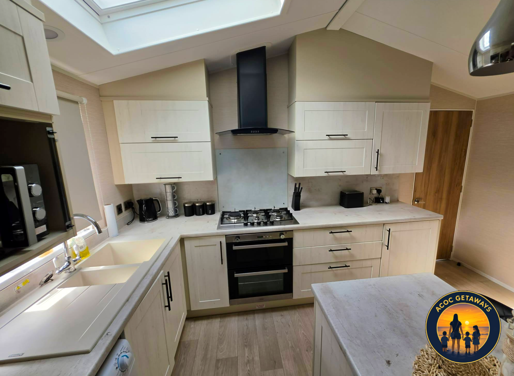 A modern kitchen with white cabinets, a black range hood, a gas stove, and various small appliances on the countertop. There is a window above the sink and a wooden door to the right.