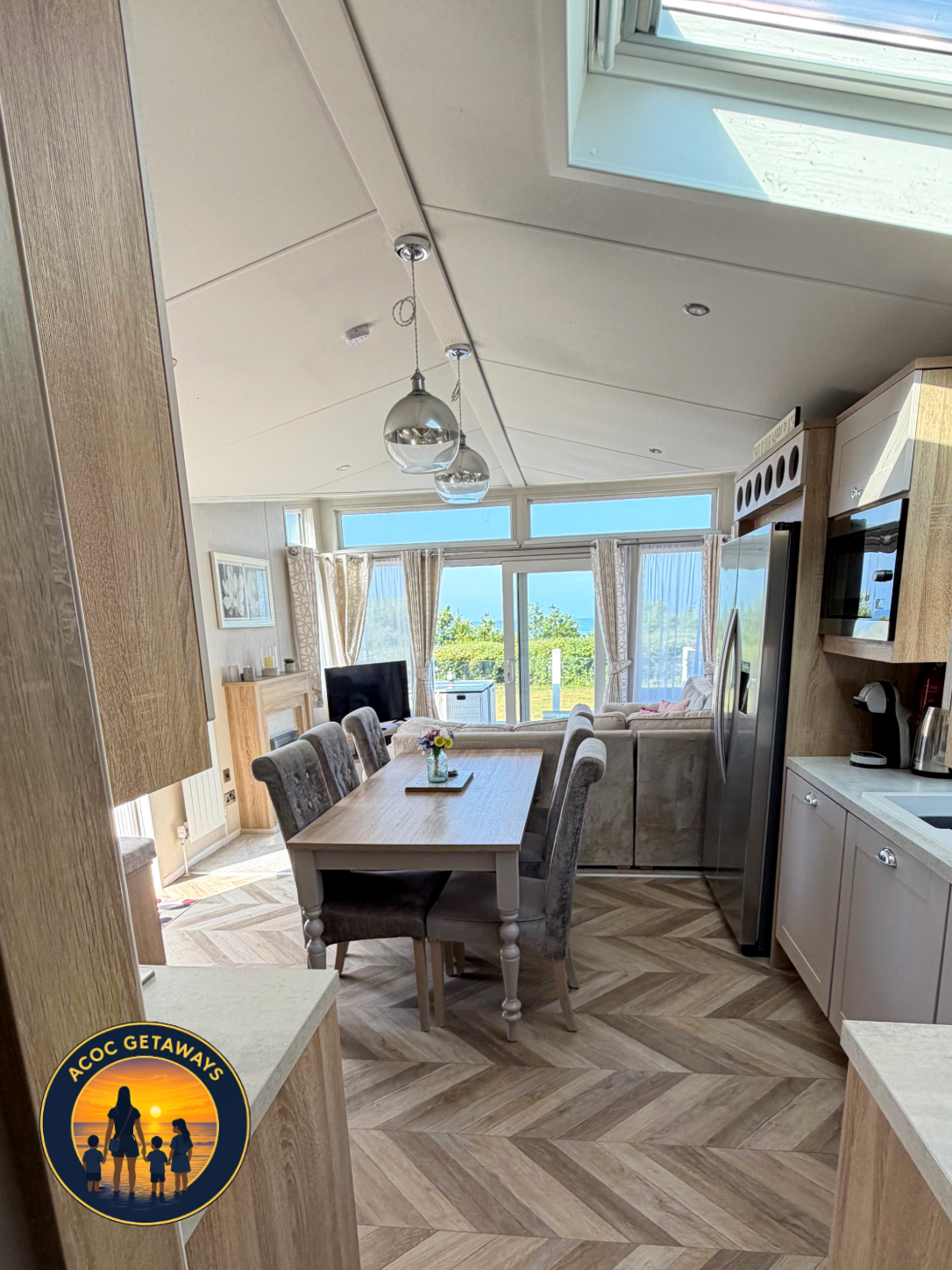 Bright, open kitchen and dining area with large windows and sliding glass doors showing a sunny outdoor view, featuring a wooden dining table with upholstered chairs, a flat-screen TV, and modern appliances.
