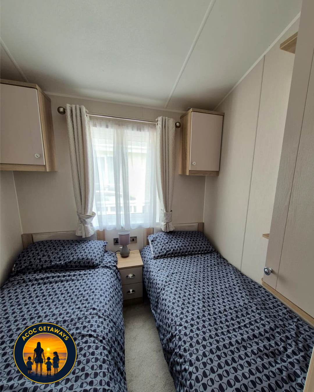 A cozy bedroom with two single beds with geometric patterned blue and black bedding, a small nightstand with a lamp, a window with sheer white curtains, and built-in cabinets on the walls.