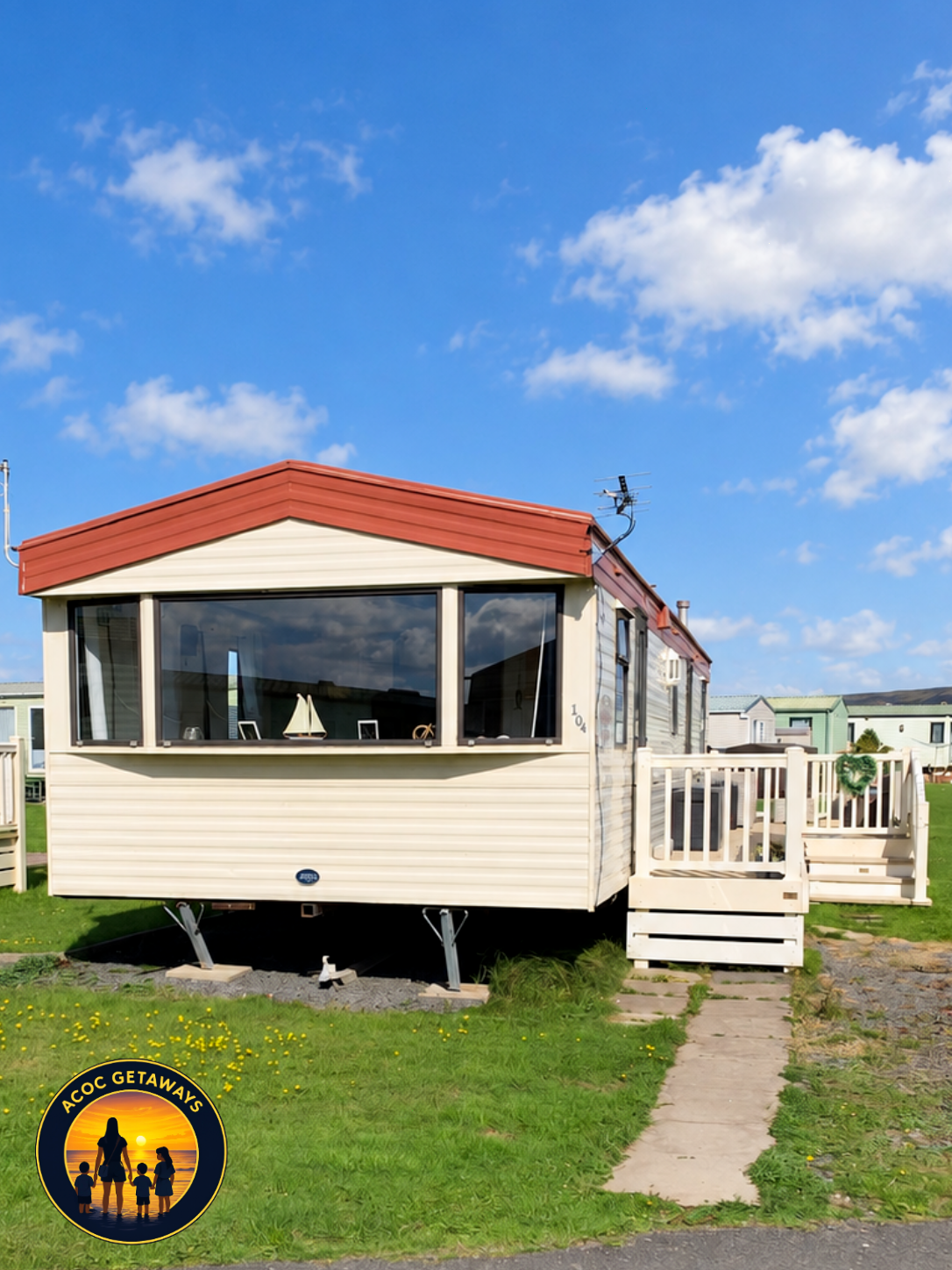A beige mobile home with a red roof and large front windows, situated on a grassy area with a stone pathway in front. The sky is blue with white clouds and other mobile homes are visible in the background.