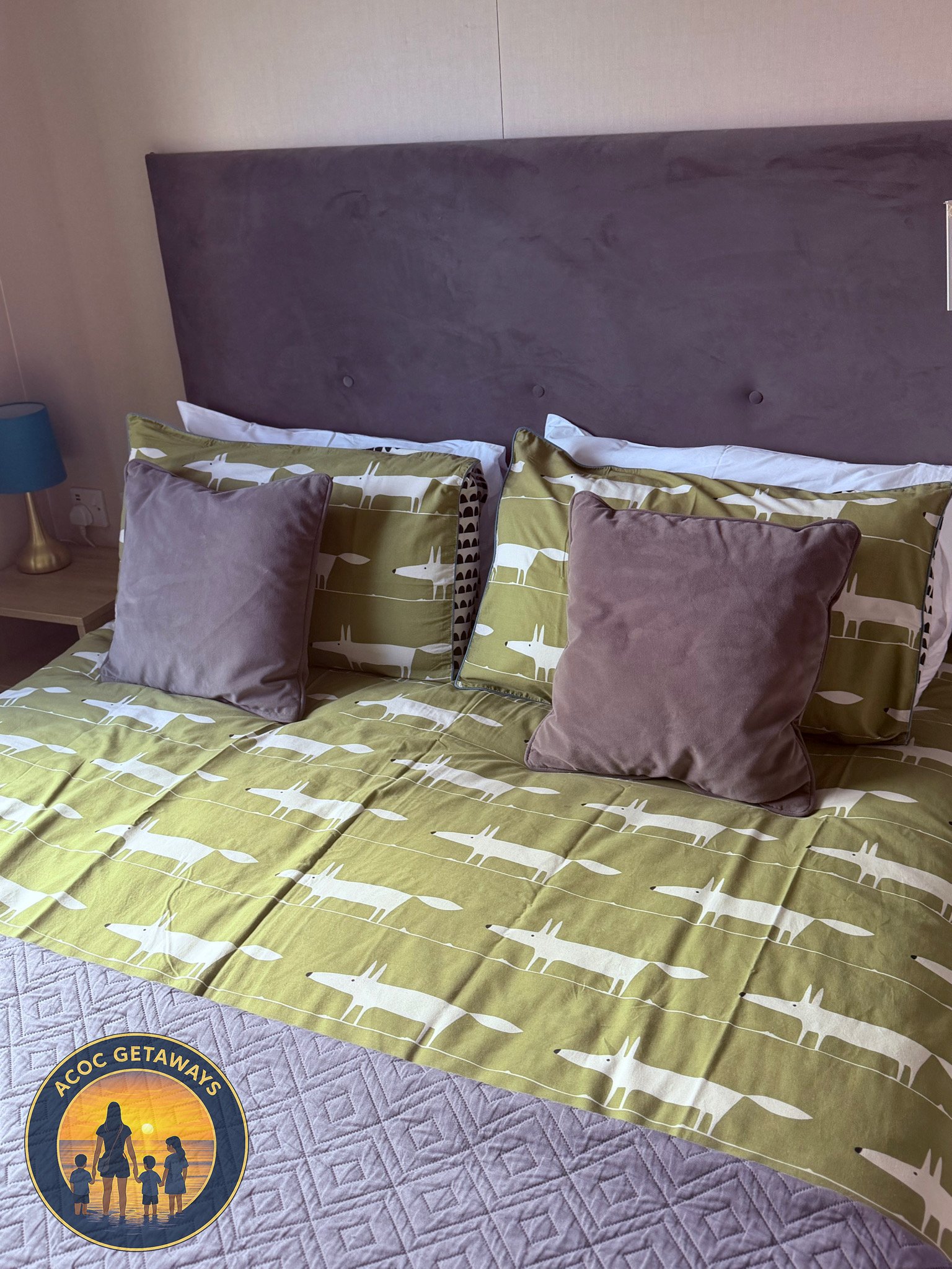 A bed with green and white dinosaur patterned bedding, two brown pillows, and two standard pillows with white pillowcases, in a room with a beige headboard and a table lamp on the nightstand.
