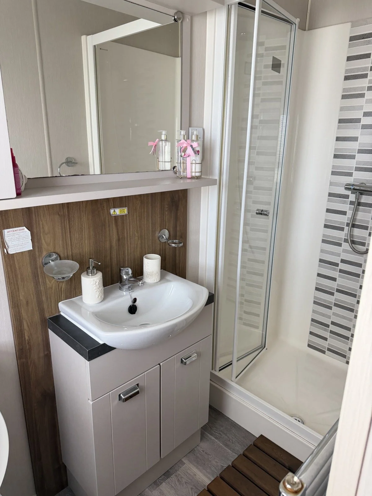 A small bathroom with a white sink, mirror, and cabinet, next to a glass-enclosed shower with gray tile accents.