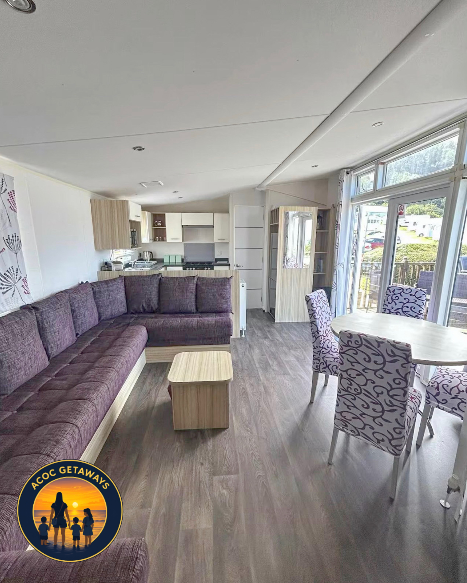 Interior of a room with a large purple sectional sofa, a light wood coffee table, and four patterned chairs around a white round table. The room has large windows with curtains, a view of a porch deck outside, and a small kitchen area in the backgrou