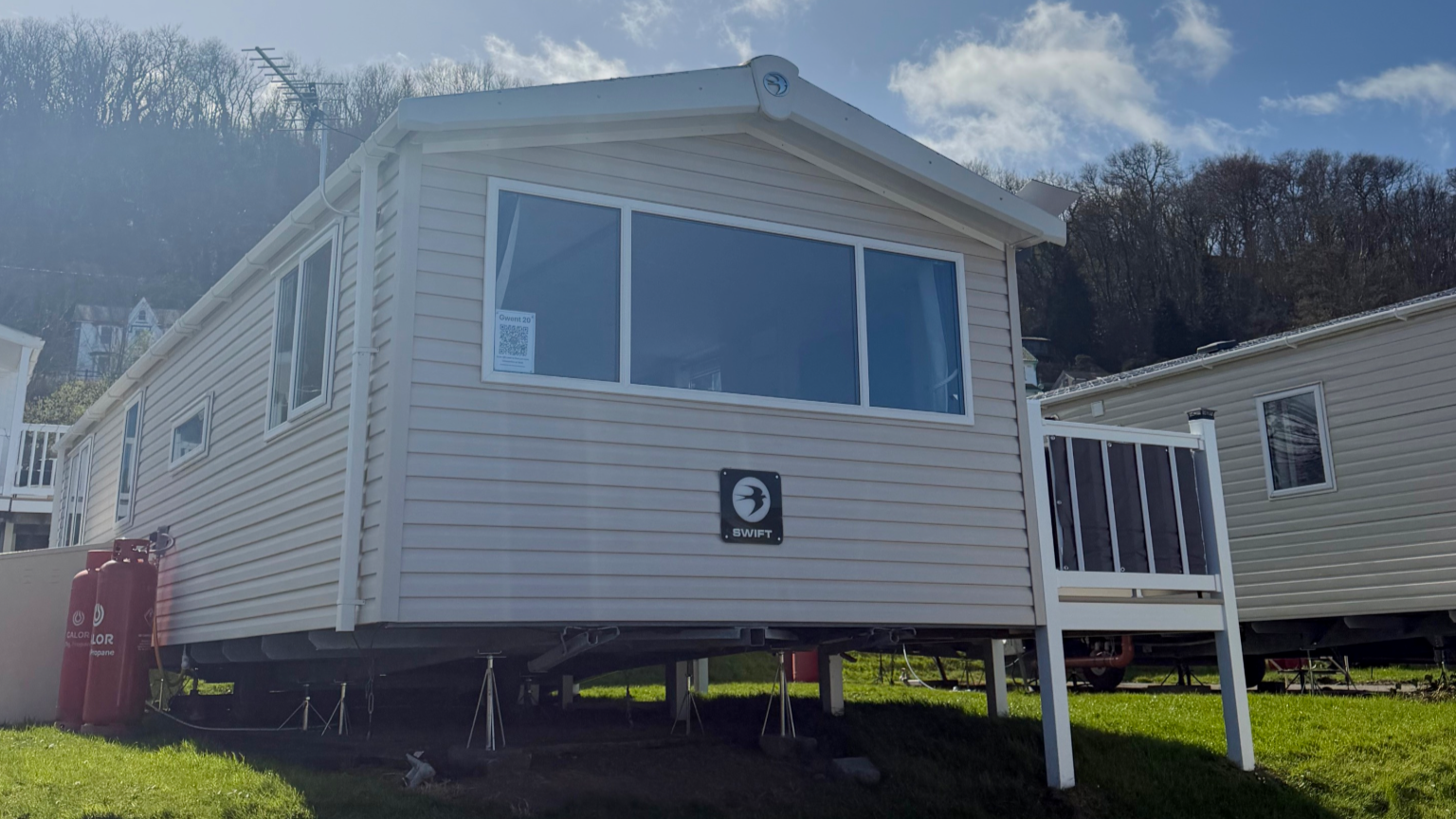 A white mobile home elevated on supports with a porch, surrounded by grass, with propane tanks nearby, under a partly cloudy sky.