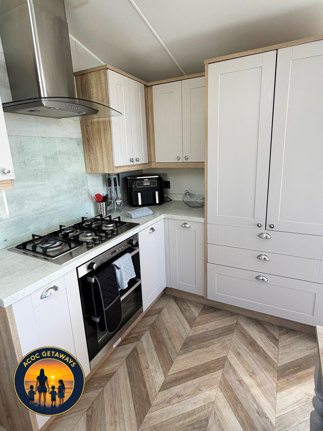 A modern kitchen with white and wood cabinets, gas stove, oven, black air fryer, and a small countertop with utensils, a towel, and a wire basket, with a herringbone wood-look floor.