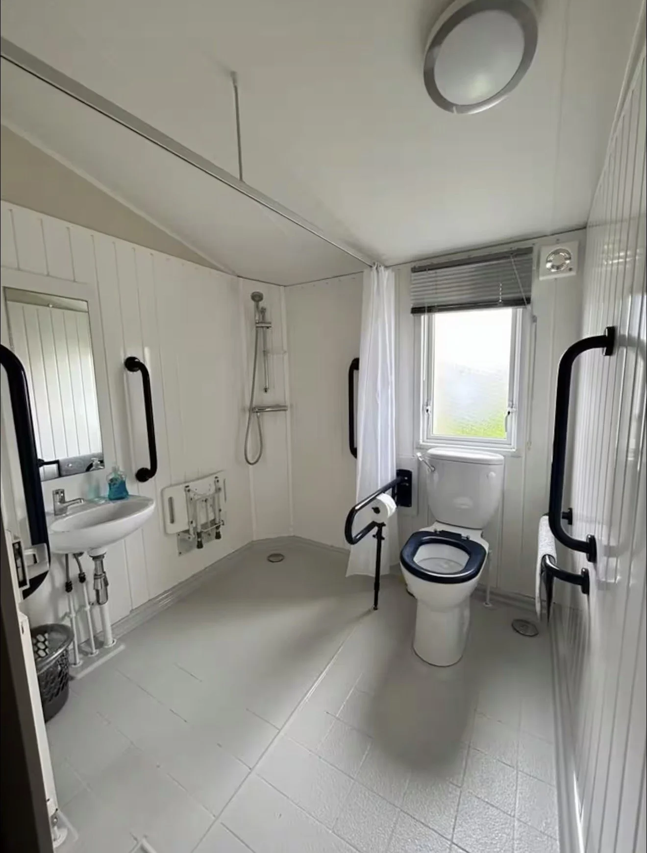 Accessible bathroom with white paneling, grab bars, a shower with a handheld showerhead, a small sink, and a toilet, all designed for wheelchair use. There are two small mirrors, a window with blinds, and a ceiling-mounted light fixture.