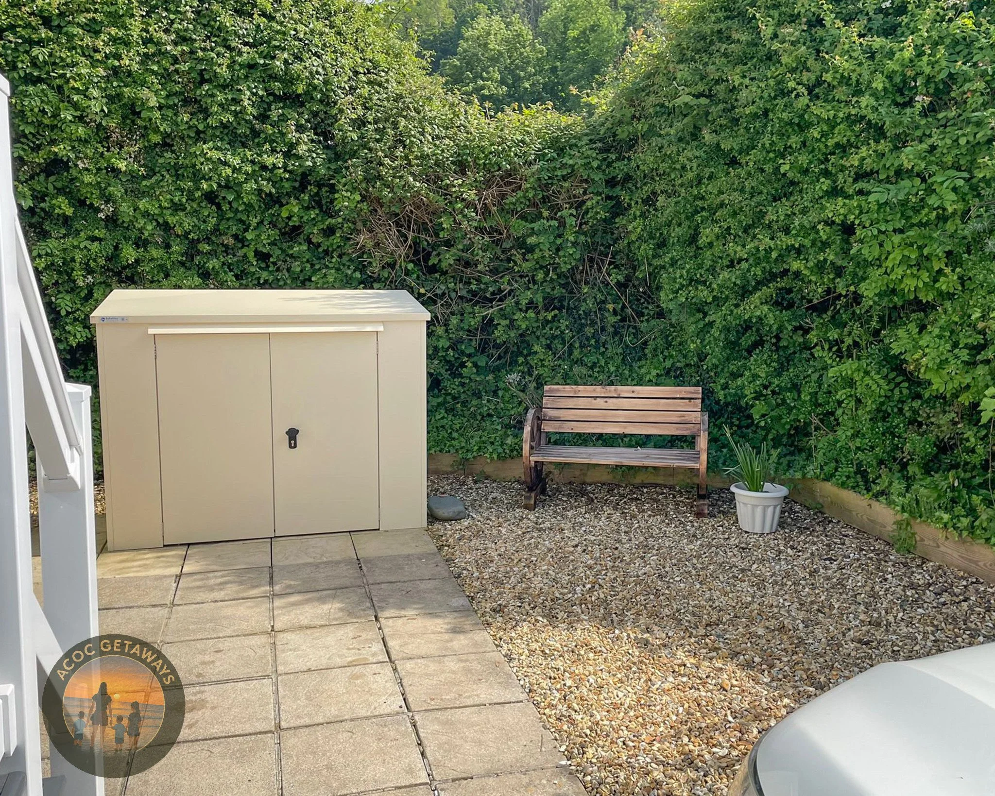 Outdoor backyard area with a beige storage shed, a wooden bench, a potted plant, and a gravel ground, surrounded by green bushes.