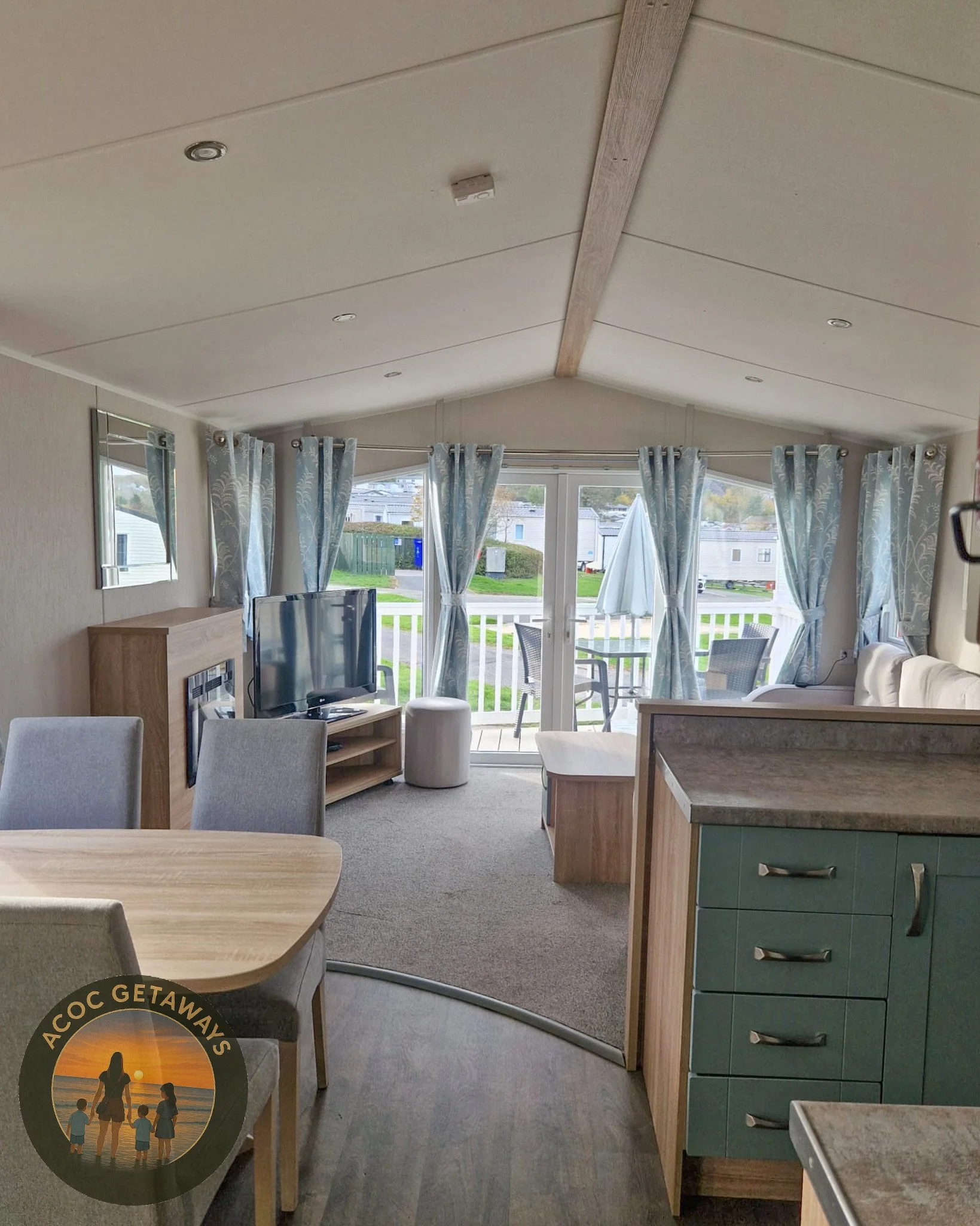 Interior of a living room with a dining area, beige carpet, light wood furniture, blue curtains at a sliding glass door leading to a balcony with outdoor chairs and an umbrella, view of a mobile home park outside.
