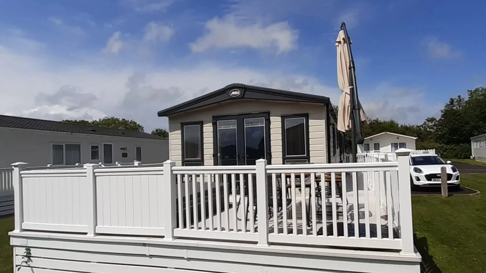 Rear view of a modern tiny house on a deck with white railing, outdoor patio furniture, large parasol, parked white car, neighboring houses, green lawn, and partly cloudy blue sky.