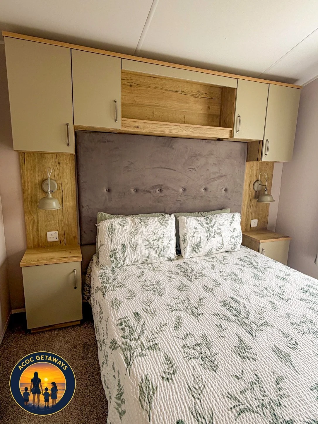 A bedroom with a double bed featuring a green leaf patterned white quilt and matching pillows, a tall grey upholstered headboard, built-in wooden and beige cabinets above and beside the bed, two beige wall-mounted reading lamps, and beige bedside cab