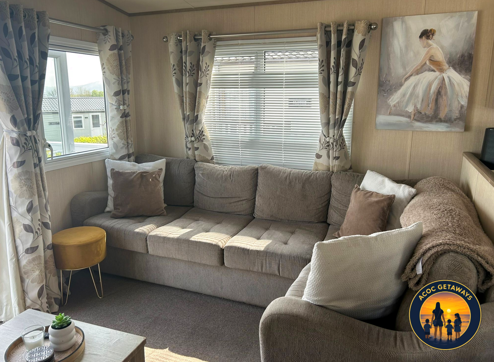 A cozy living room corner with a beige couch adorned with various cushions, next to a window with blinds, and floral curtain panels. There's a small round yellow side table nearby, and a coffee table with a succulent plant and candles in the foregrou