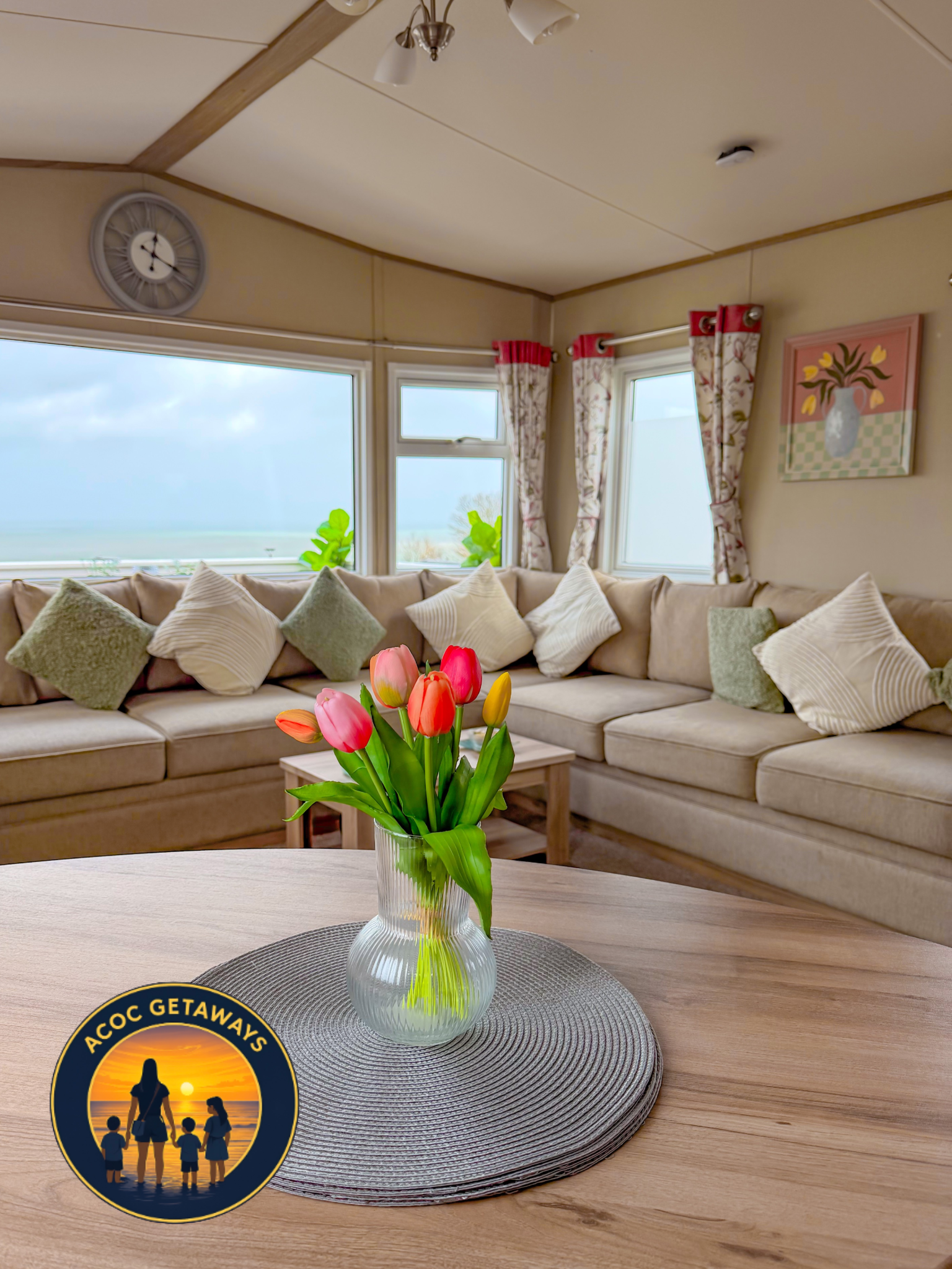 A cozy living room with a beige sectional sofa filled with white and pale green pillows, a round wooden coffee table, and a small side table. A vase of pink, red, yellow, and orange tulips is on the coffee table. There are windows with floral curtain