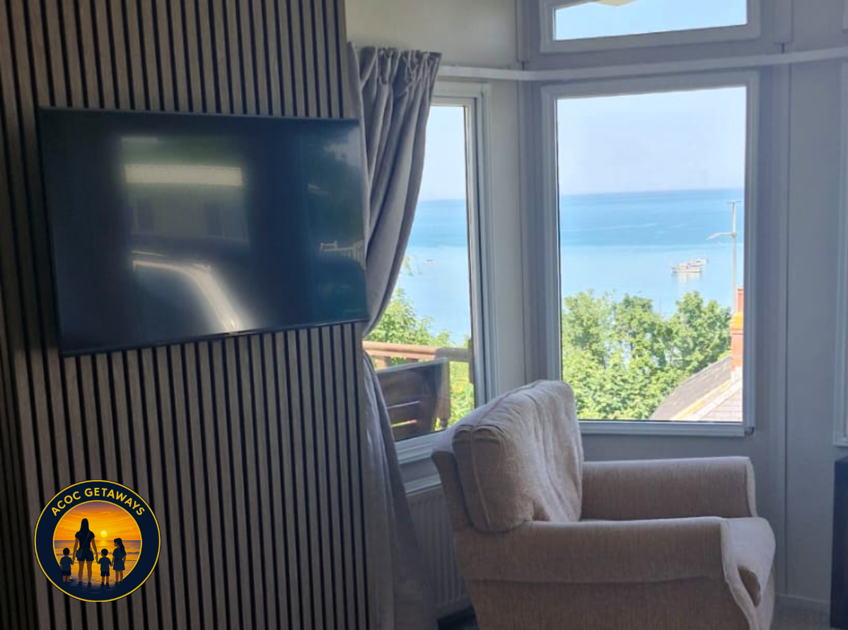 Room with a large window overlooking a body of water with boats, a beige armchair, and a wall-mounted flat-screen TV.