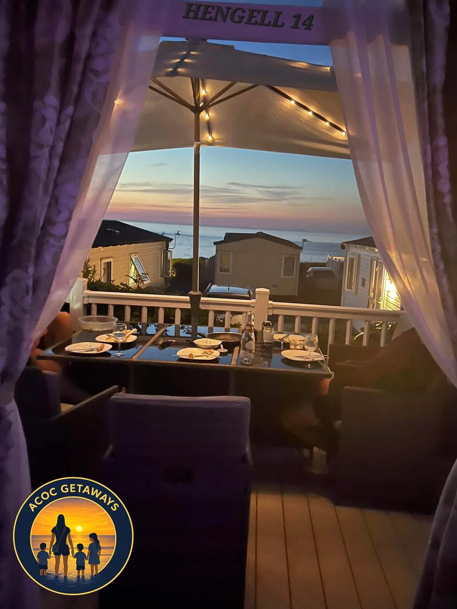 A dining table set for four with plates, glasses, and condiments, seen through a curtained doorway overlooking a balcony with an ocean view during sunset, with a large umbrella and neighboring houses in the background.