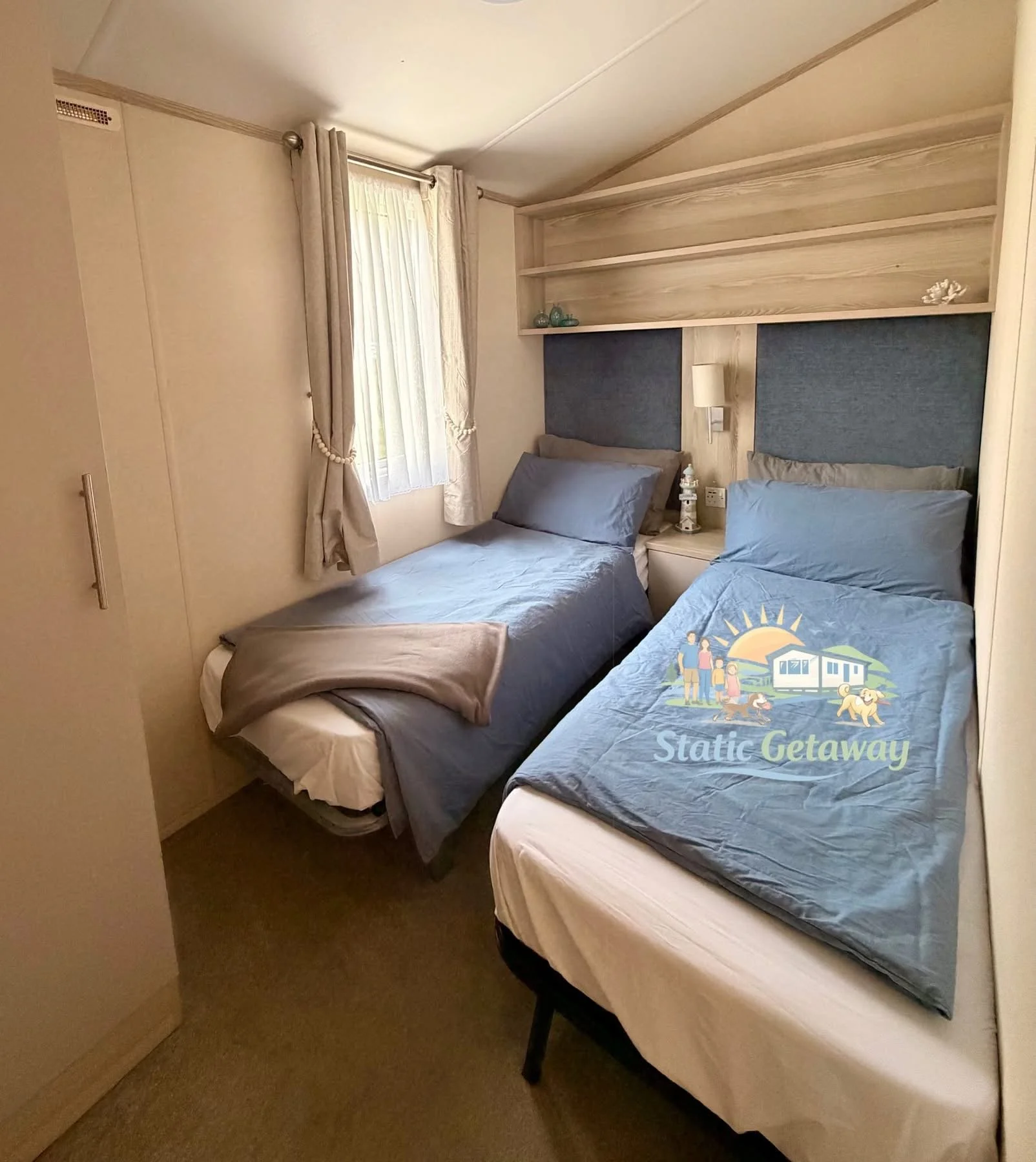 Bedroom with two single beds, blue and gray bedding, a nightstand with a lighthouse lamp between the beds, a window with white curtains, and a built-in closet with open shelving above the beds in a cozy, light-colored room.