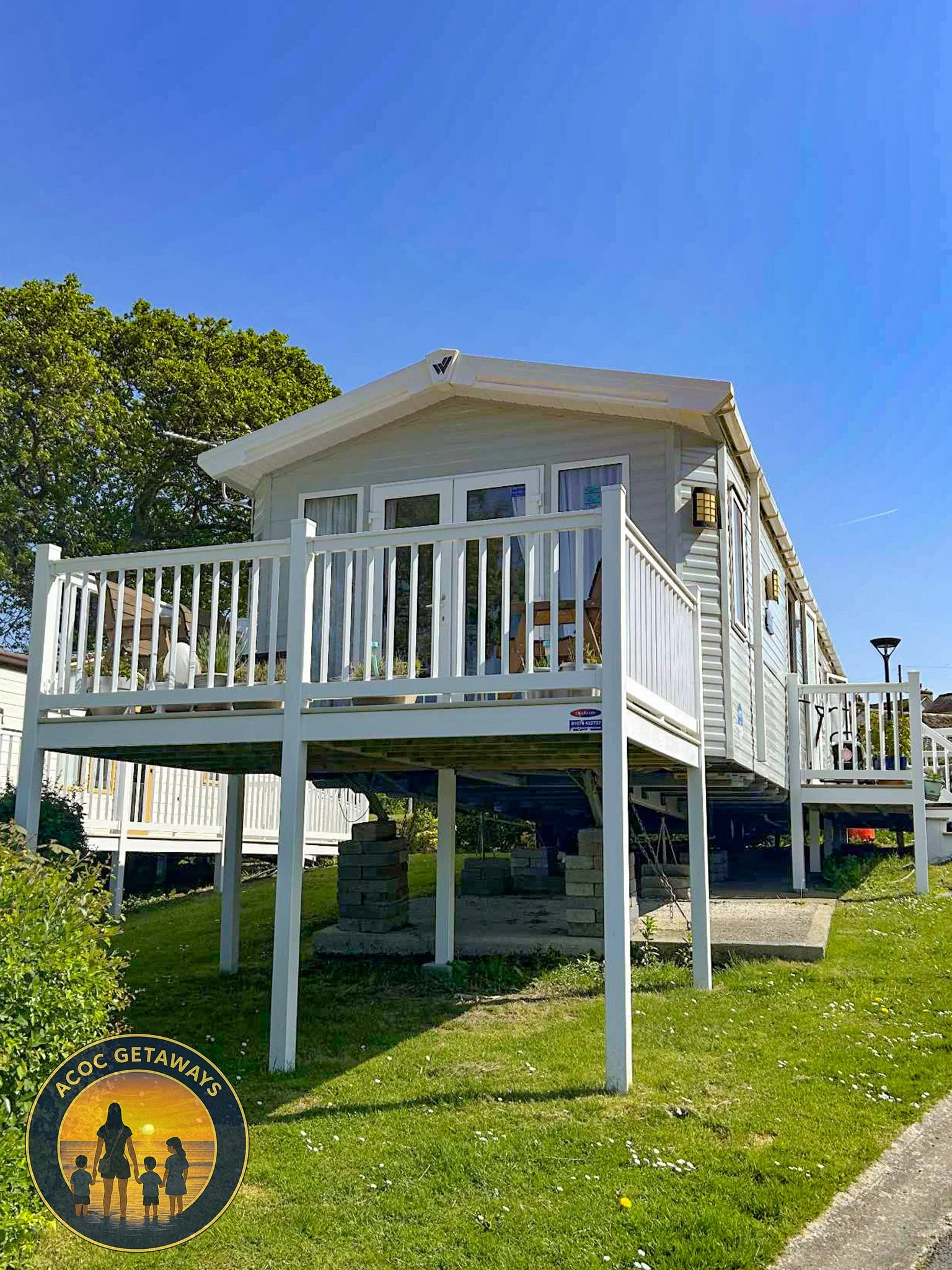 A house built on stilts with a white wooden deck and railing, situated on a grassy lawn under a clear blue sky. The house has gray siding and multiple windows.
