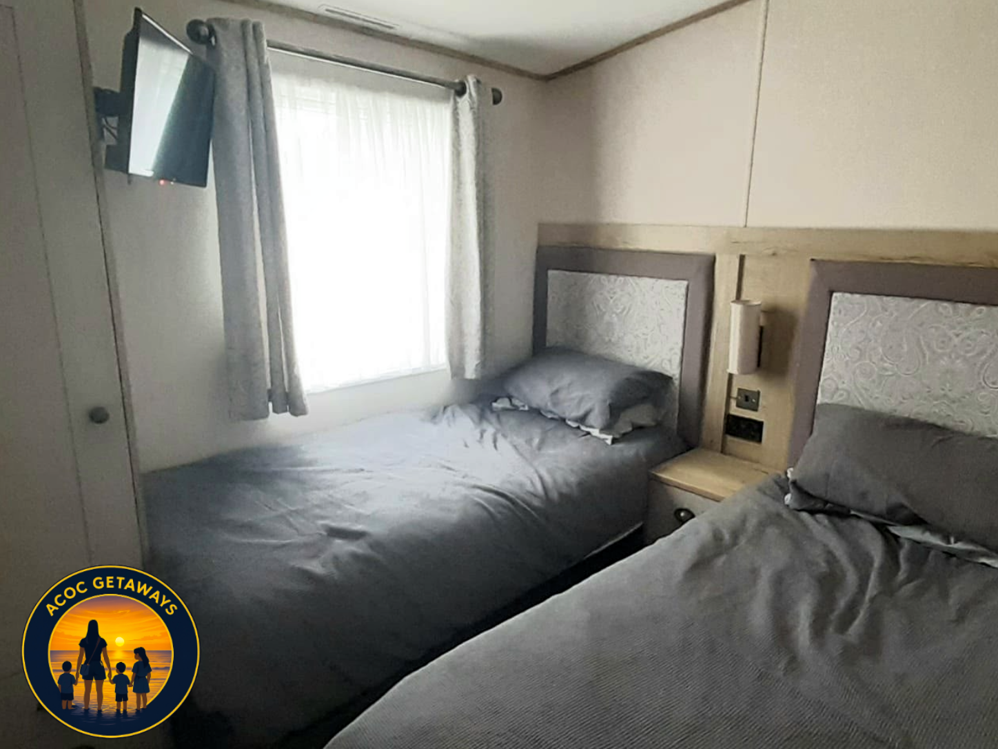 A hotel room with two single beds covered in gray bedding, a window with light curtains, a small wall-mounted TV, and a nightstand between the beds with electrical outlets and a wall-mounted reading light.