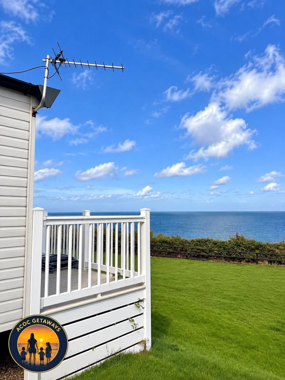 A backyard with green grass, a white wooden fence, a house with beige siding, and an ocean view in the distance under a partly cloudy sky.
