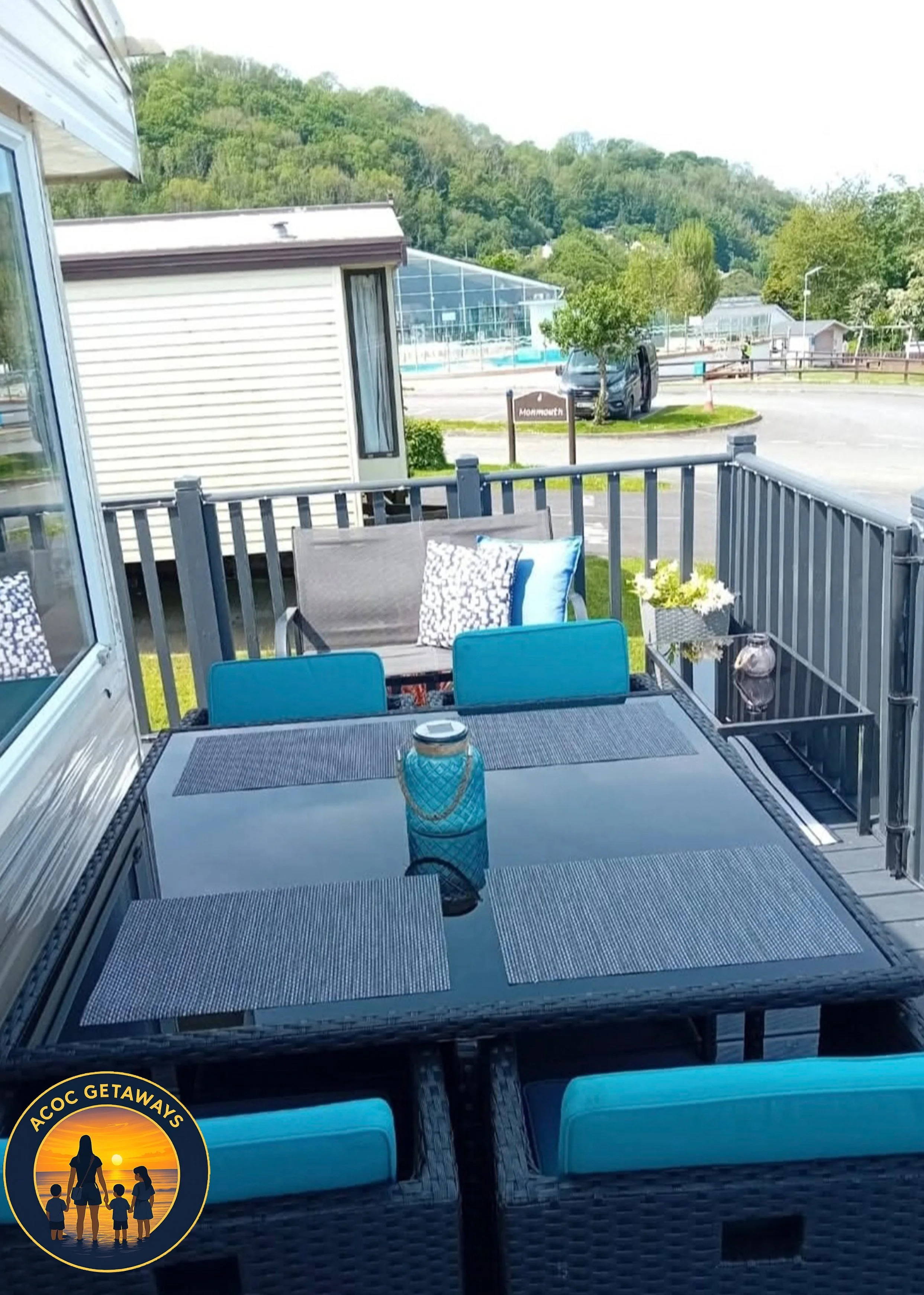 Outdoor patio with a glass table, six blue chairs, and a gray patio sofa with two cushions, one patterned and one plain blue. There is a small vase with flowers and a decorative blue jar on the table. The patio is enclosed by a gray railing, overlook
