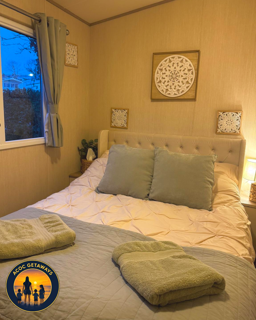 A cozy bedroom with beige walls and a window with light green curtains. The bed has a white quilted comforter, two large light green pillows, and towels on top. There are decorative wall hangings and a small bedside table with a basket and plant. A s