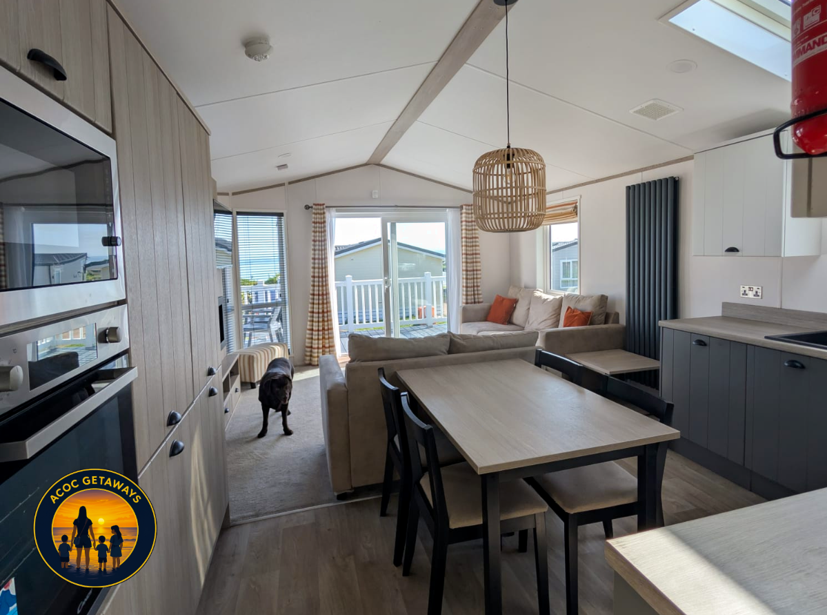 Interior of a cozy living room and kitchen area with a dining table, beige sofa with orange pillows, and large glass doors leading to a balcony with outdoor furniture. A black dog is near the sofa. Natural light fills the space, and there is a wooden