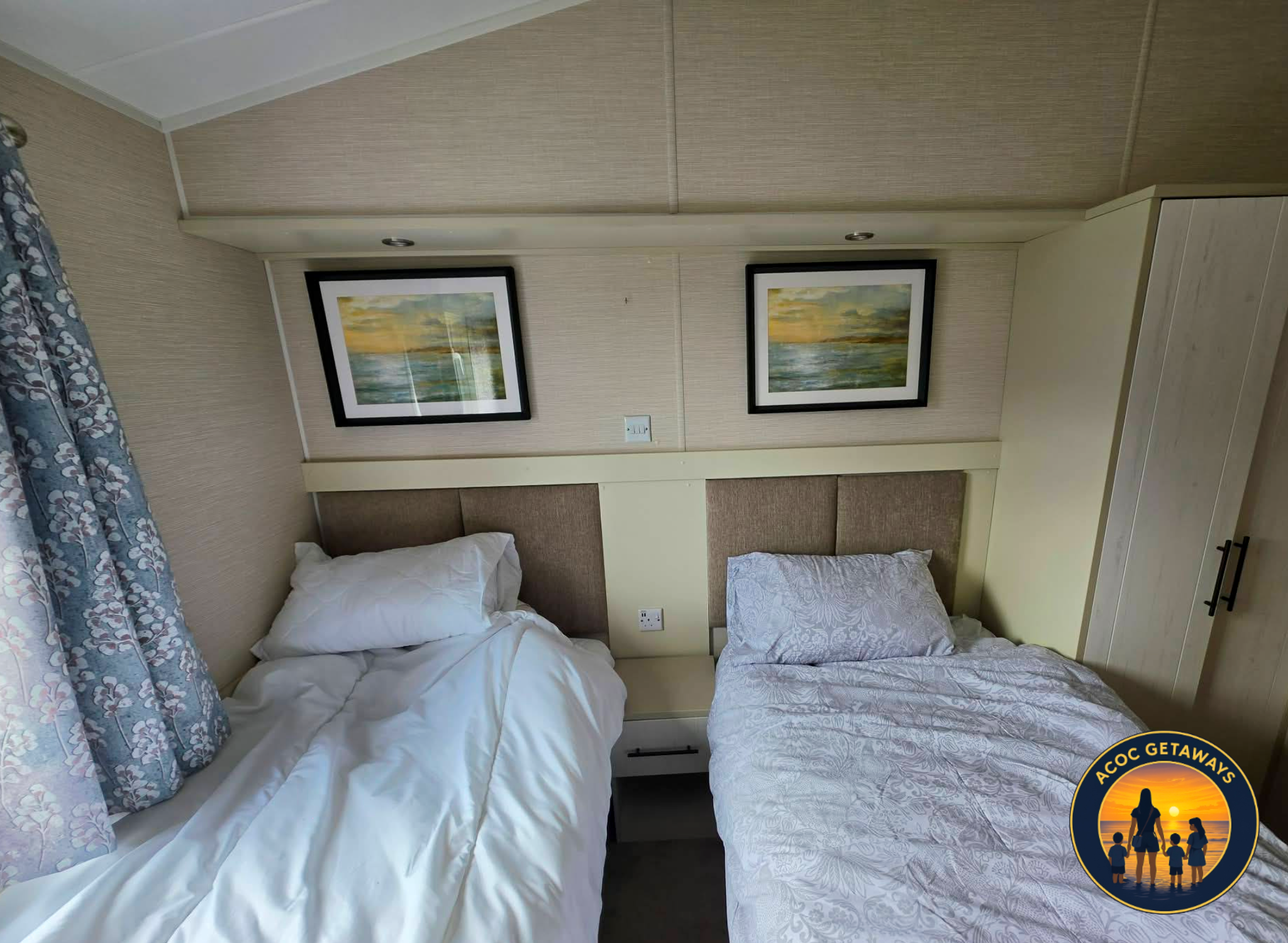 Two beds with white bedding, one with floral pillowcases, a beige headboard, and framed landscape paintings above. A cream-colored wall with two small lights and a white electrical outlet.