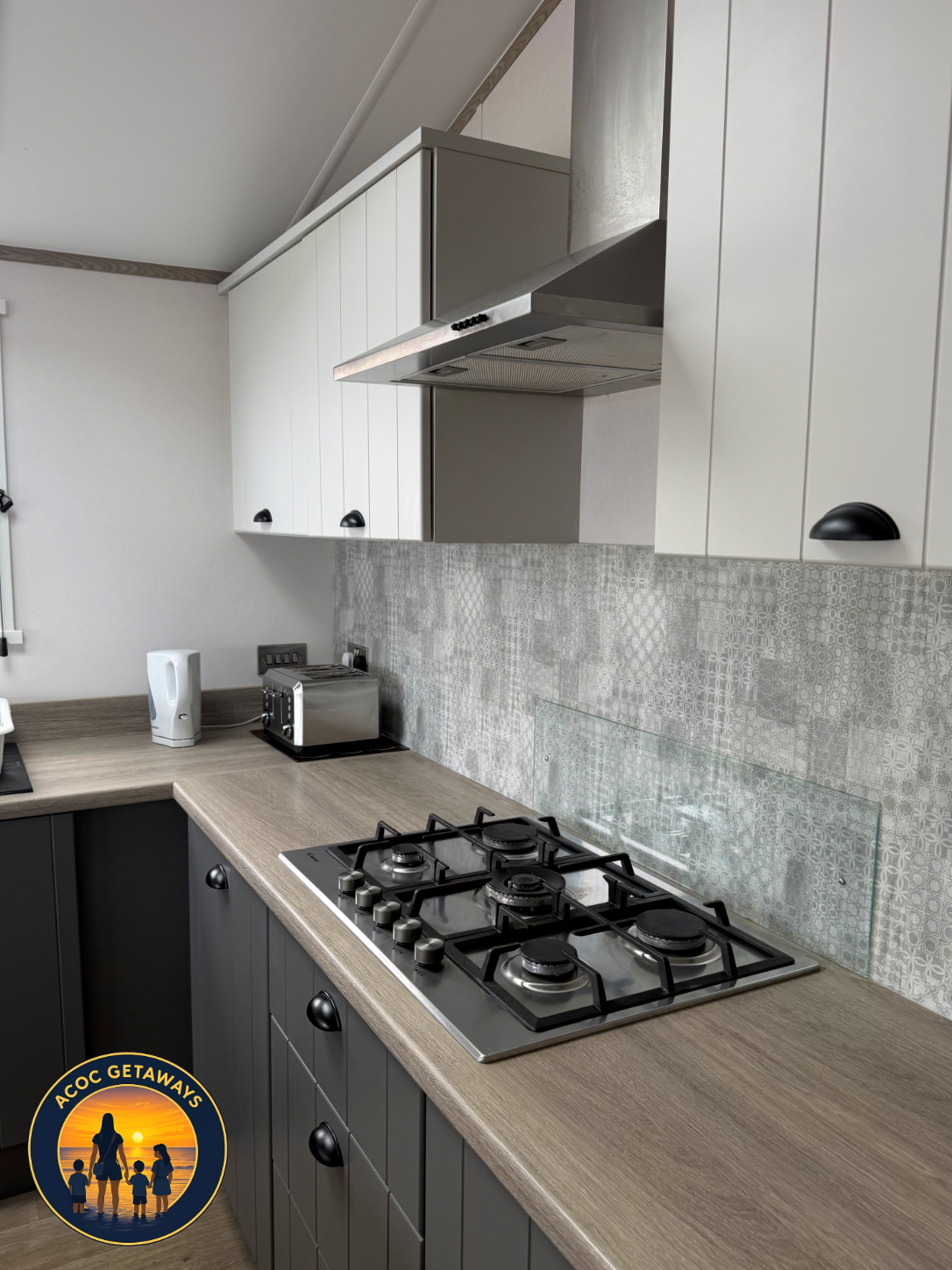 Modern kitchen with dark lower cabinets, light countertop, gas stove, and white upper cabinets. Includes toaster, electric kettle, and patterned backsplash. Logo in bottom left corner: "AOC GETAWAYS" with silhouette of a woman and children at sunset.
