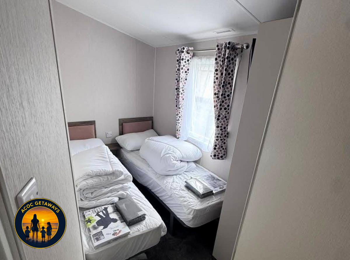 A small bedroom with two single beds, one with folded bedding and magazines on top, the other with bedding and a pillow. A window with polka dot curtains lets in natural light. The room has plain light-colored walls and a ceiling with a vent.