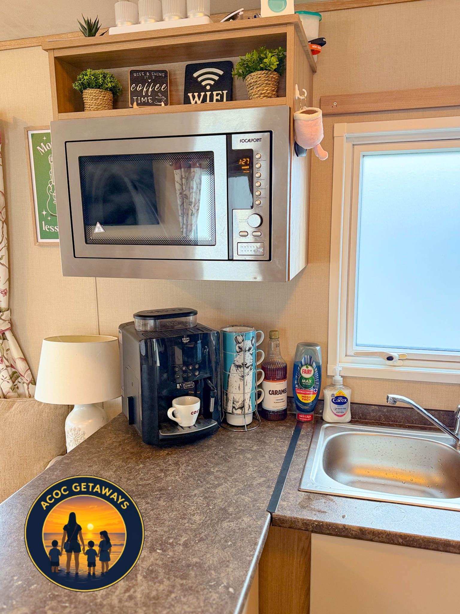 A coffee station with a microwave oven mounted above a countertop. On the counter, there is a coffee machine with a cup in it, a mug, and a stick of stirrers. To the right, there are containers of syrup, dish soap, and a sponge near a sink. On the wa