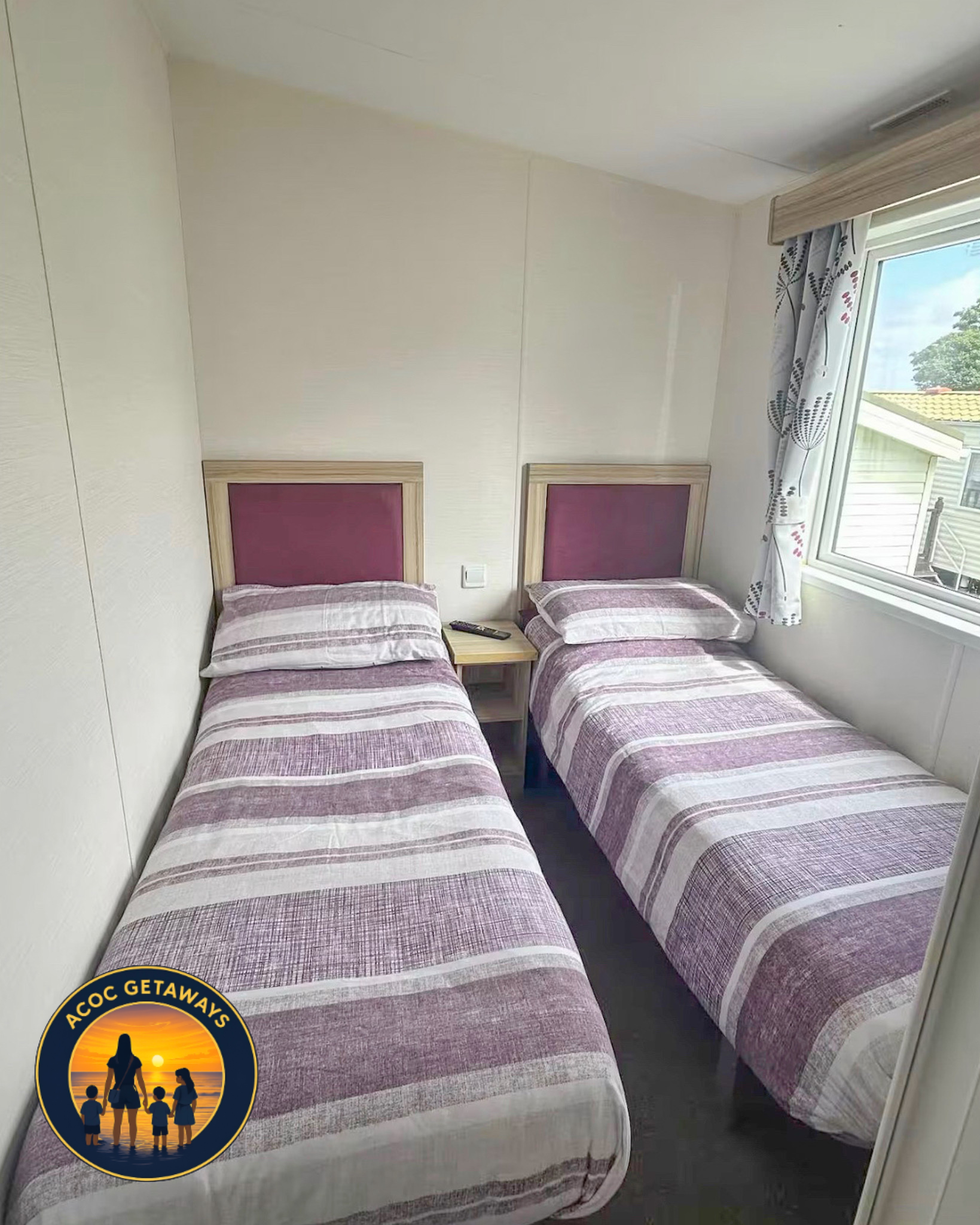 Two twin beds with purple and beige striped bedding and wooden headboards in a room with cream walls, a window with floral curtains, and a wooden window frame.