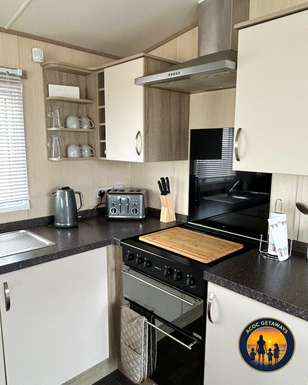 Kitchen with black countertops, white cabinets, a stainless steel stove with a wooden cutting board on top, a stainless steel kettle, a toaster, knife set, and a tea towel hanging on the oven handle. Open shelves with cups and glasses, window with bl