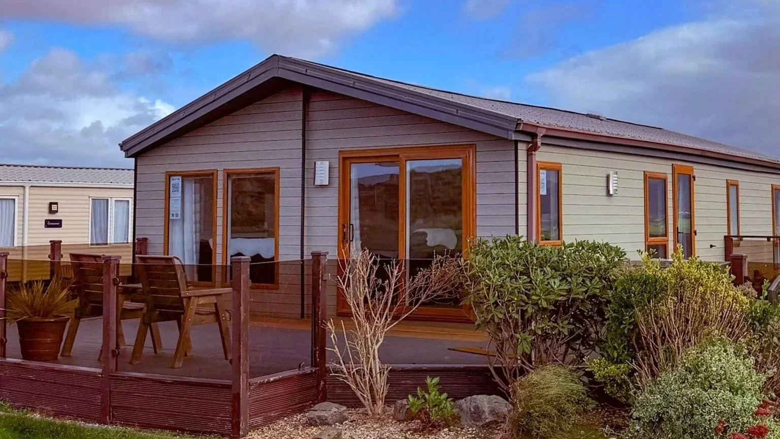 A wooden house with large glass doors and windows, surrounded by a garden with various shrubs and plants. There is a deck with a wooden table and chairs, under a partly cloudy sky.